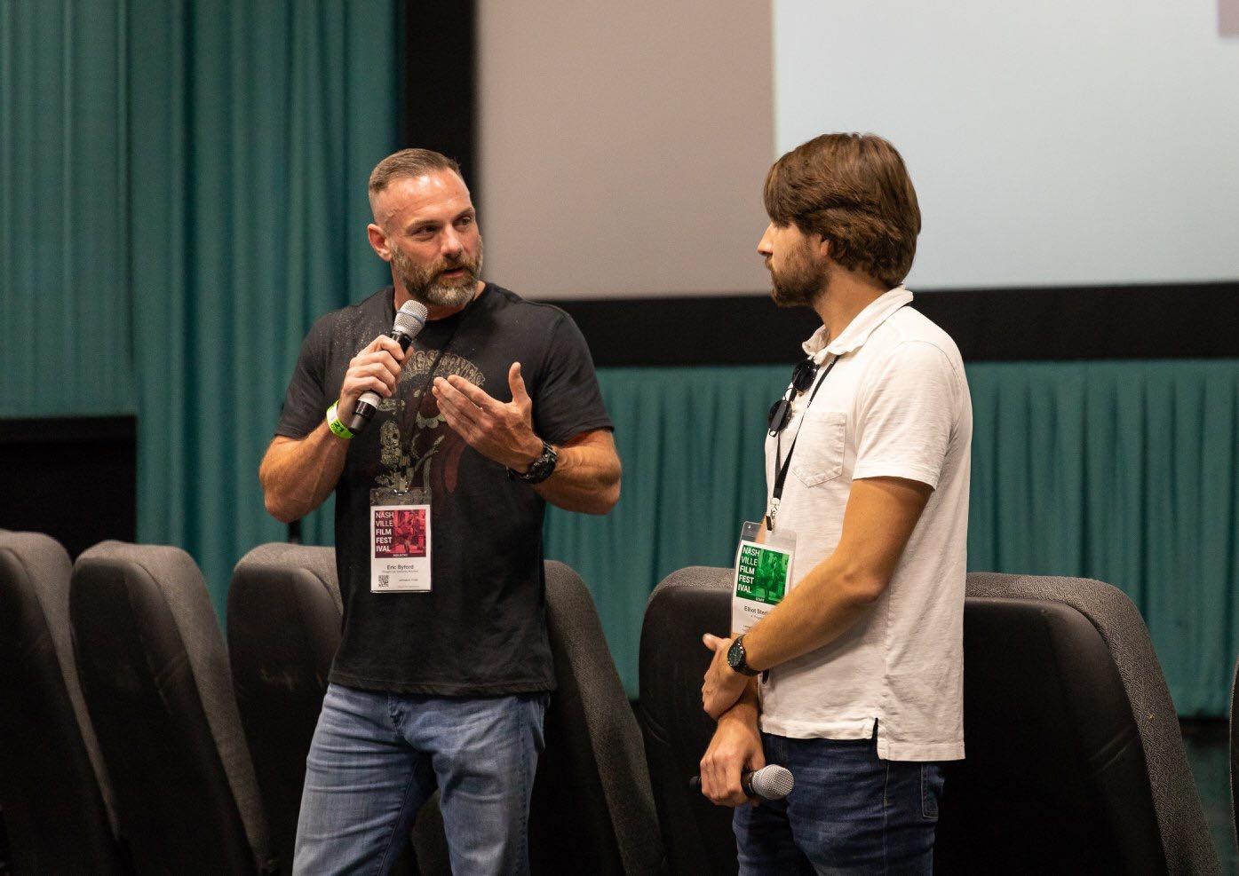 Two men are speaking on stage, one holding a microphone and gesturing, the other listening, with a large screen behind them.
