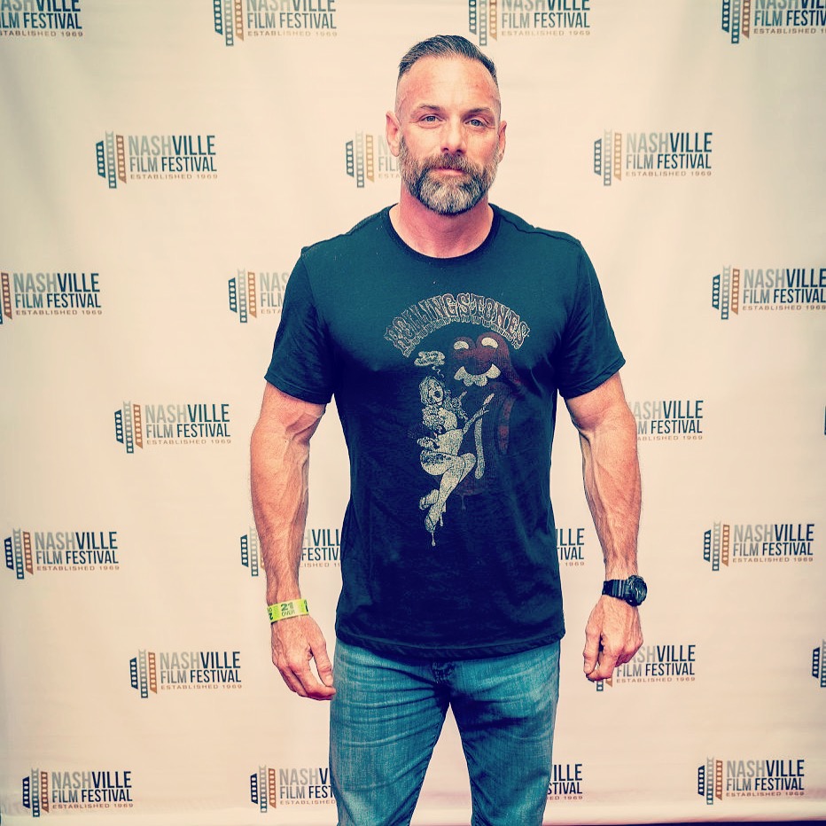 Man with beard and short hair standing in front of a Nashville Film Festival backdrop, wearing a black T-shirt and jeans.