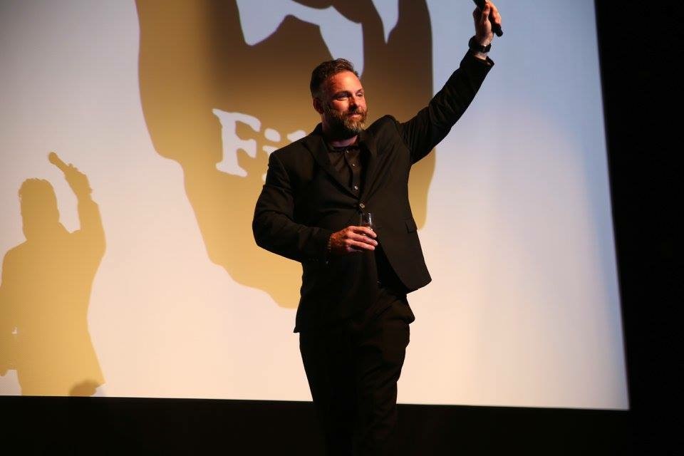 Man in dark suit holding a microphone and raising his hand on stage, with a large shadow behind him.