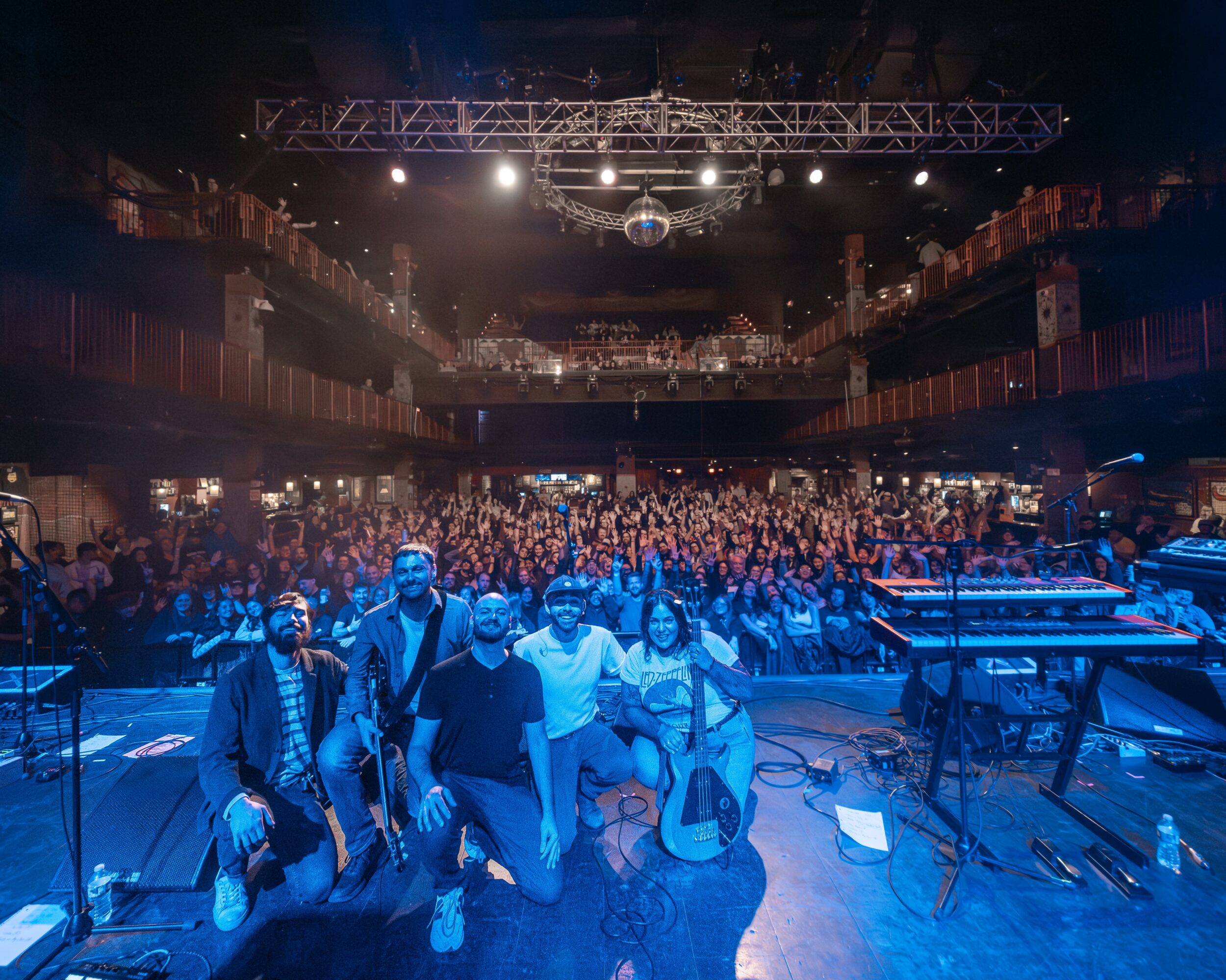 Group of people on stage in front of a large audience in a concert hall, stage equipment visible, dim lighting.