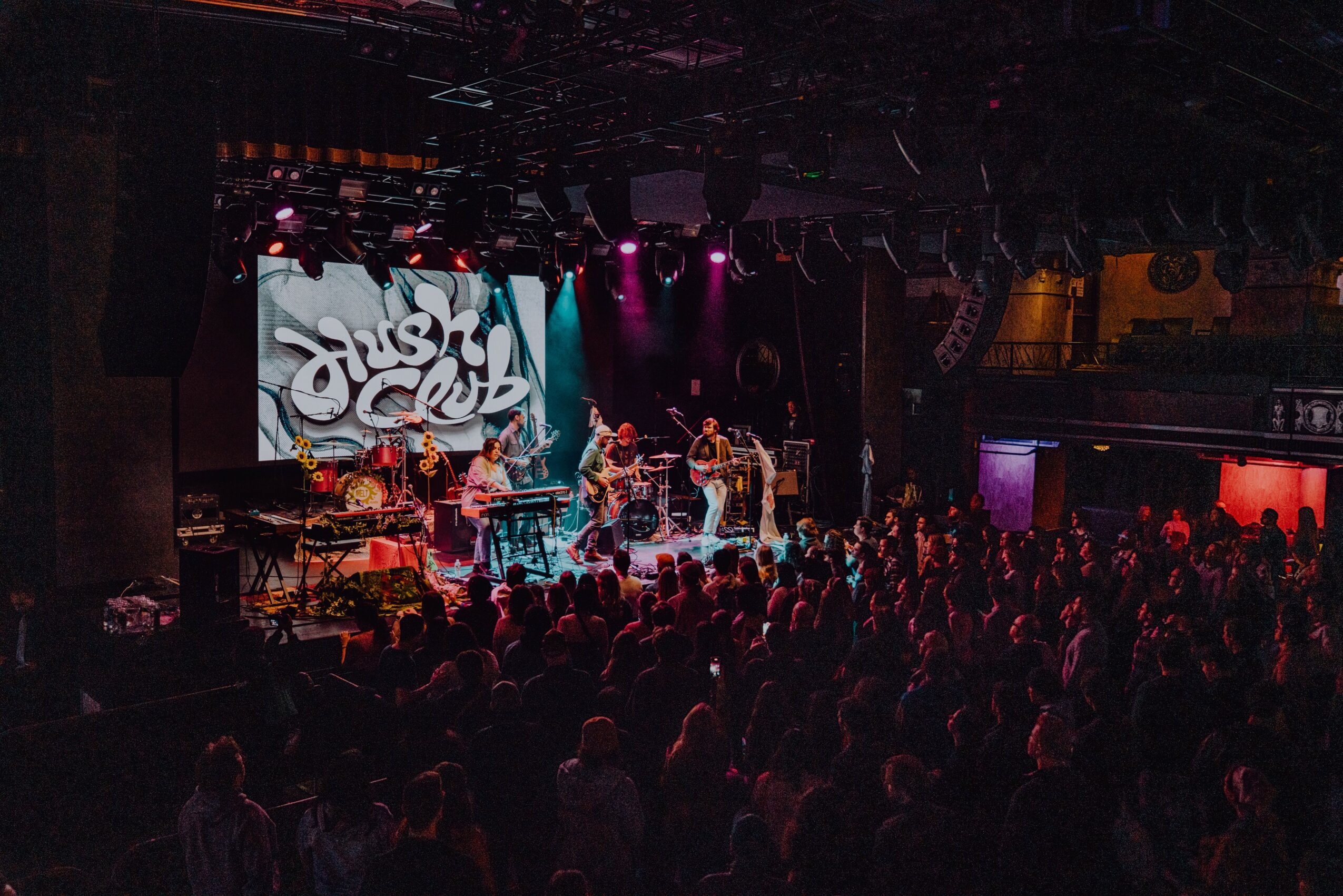 Concert stage with musicians performing, audience watching, colorful lighting, and a large screen displaying graphics.