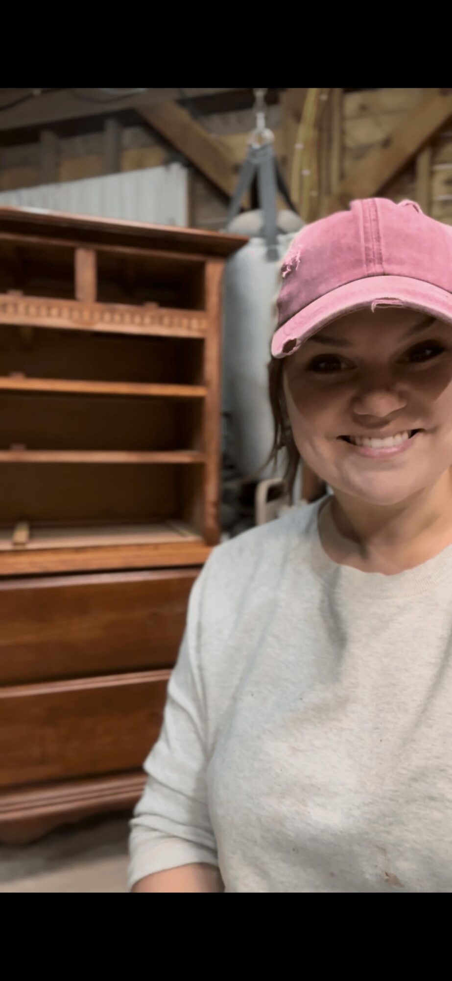 Smiling person wearing a pink cap and light-colored shirt in a workshop with wooden furniture and tools.