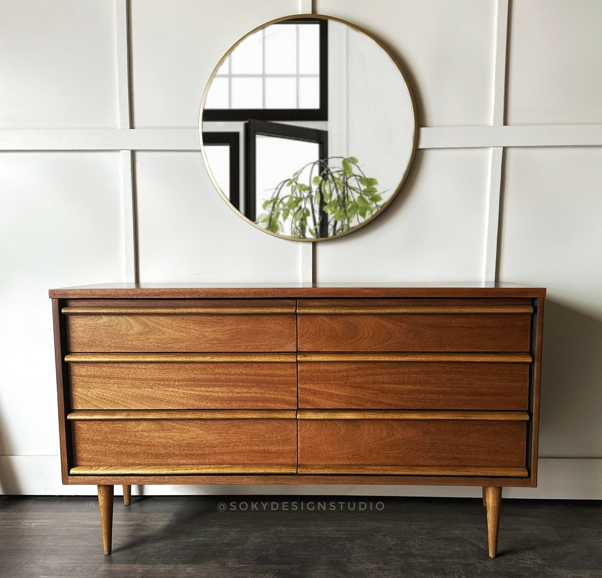 Wooden dresser with six drawers and a round mirror above it, reflecting a window and plant, against a white wall.
