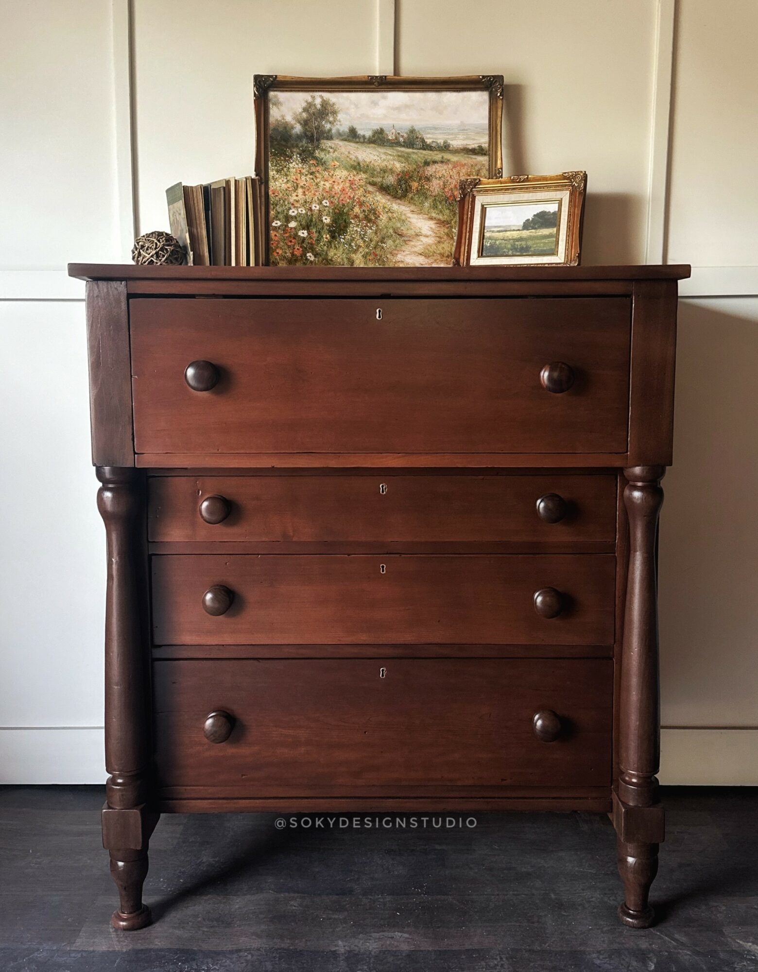 Wooden chest of drawers with books and landscape paintings on top, against a white wall.
