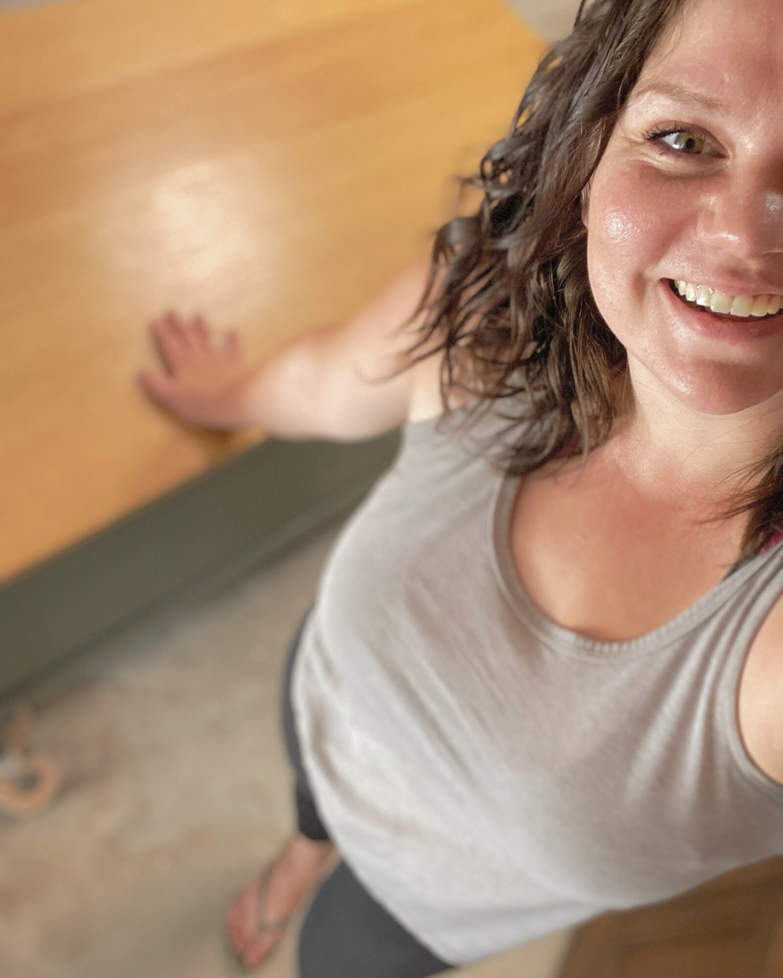 Woman smiling, taking a selfie from above, standing on a wooden floor, wearing a sleeveless top.
