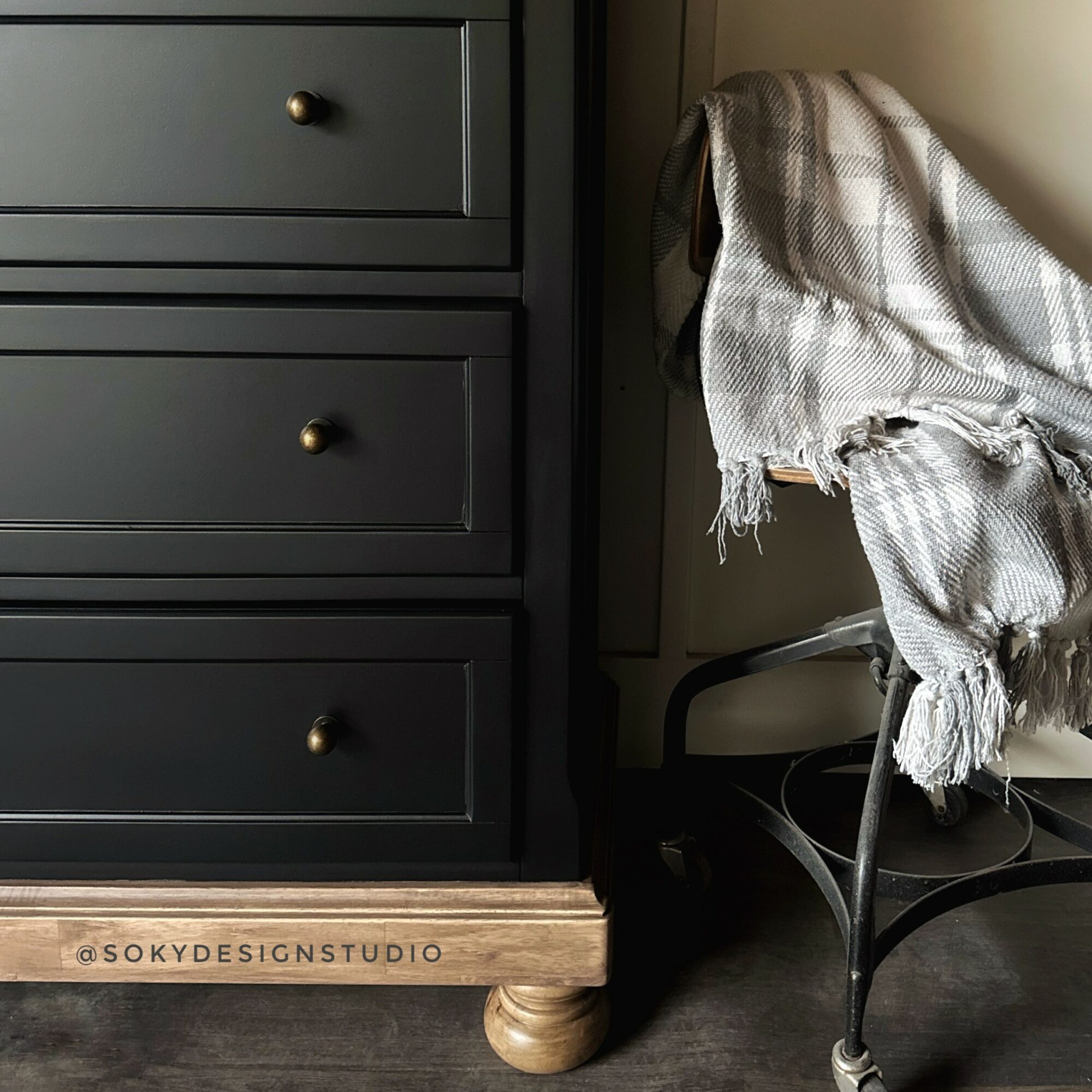 Close-up of a black dresser with three drawers and a wooden base, next to a chair with a checkered cloth draped over it, on a dark floor.