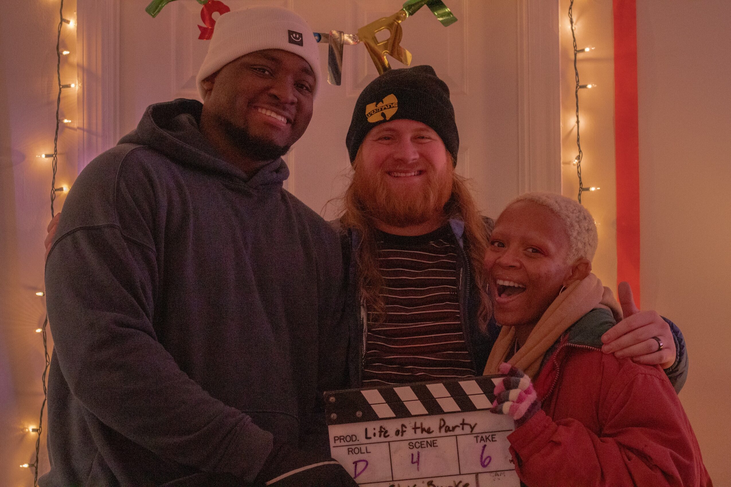 Three people smiling and hugging in front of holiday decorations, including string lights and a banner, celebrating together.