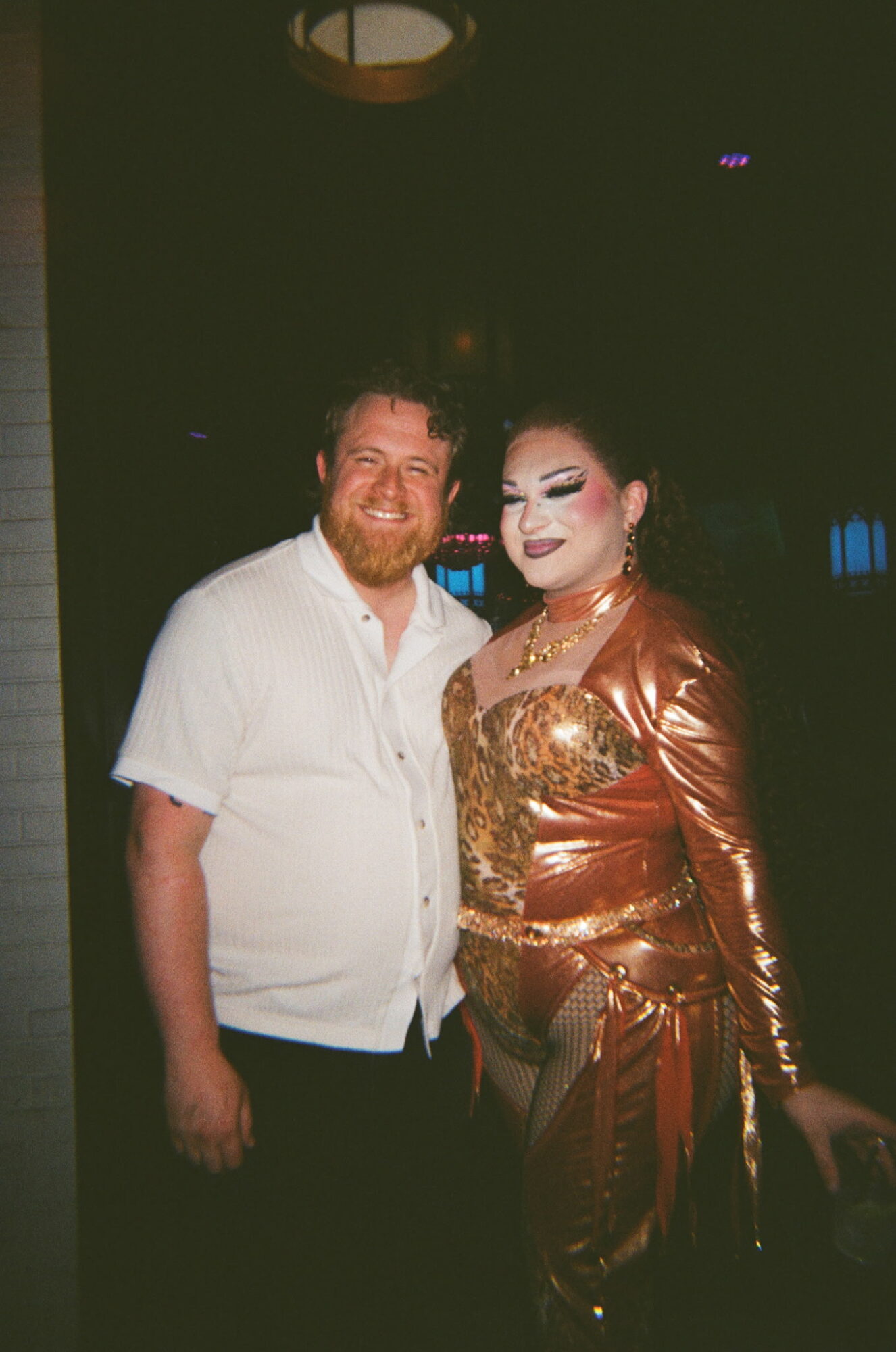 Two people, one with a beard and white shirt, and the other in elaborate costume, pose together in a dark setting.