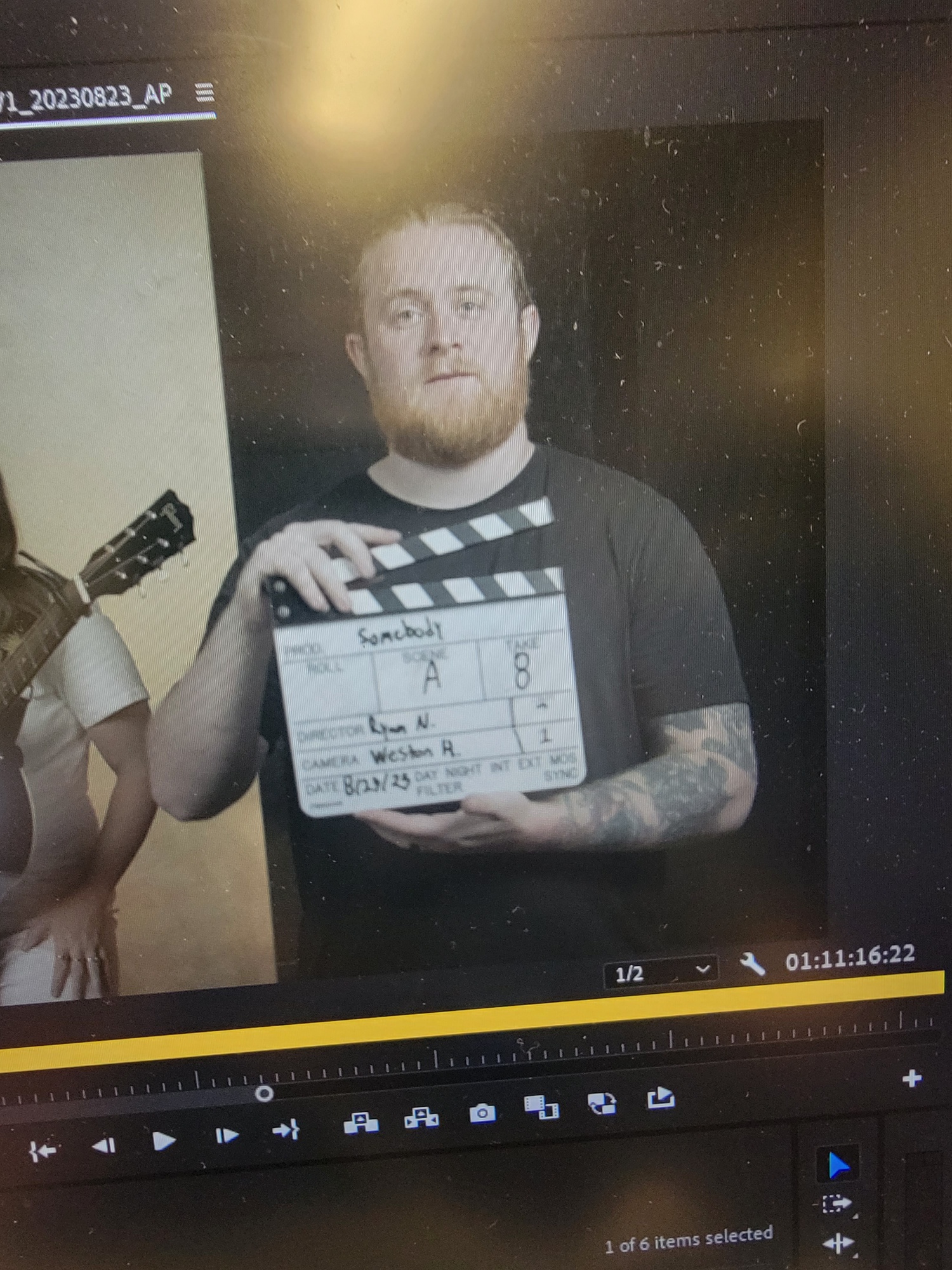 Man with tattoos holding a clapperboard, standing indoors with dark background.
