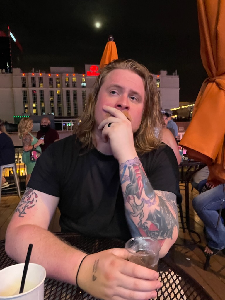 Young man with long hair and tattoos on arms, sitting outdoors at night, holding a drink, with a cityscape and umbrellas in background.