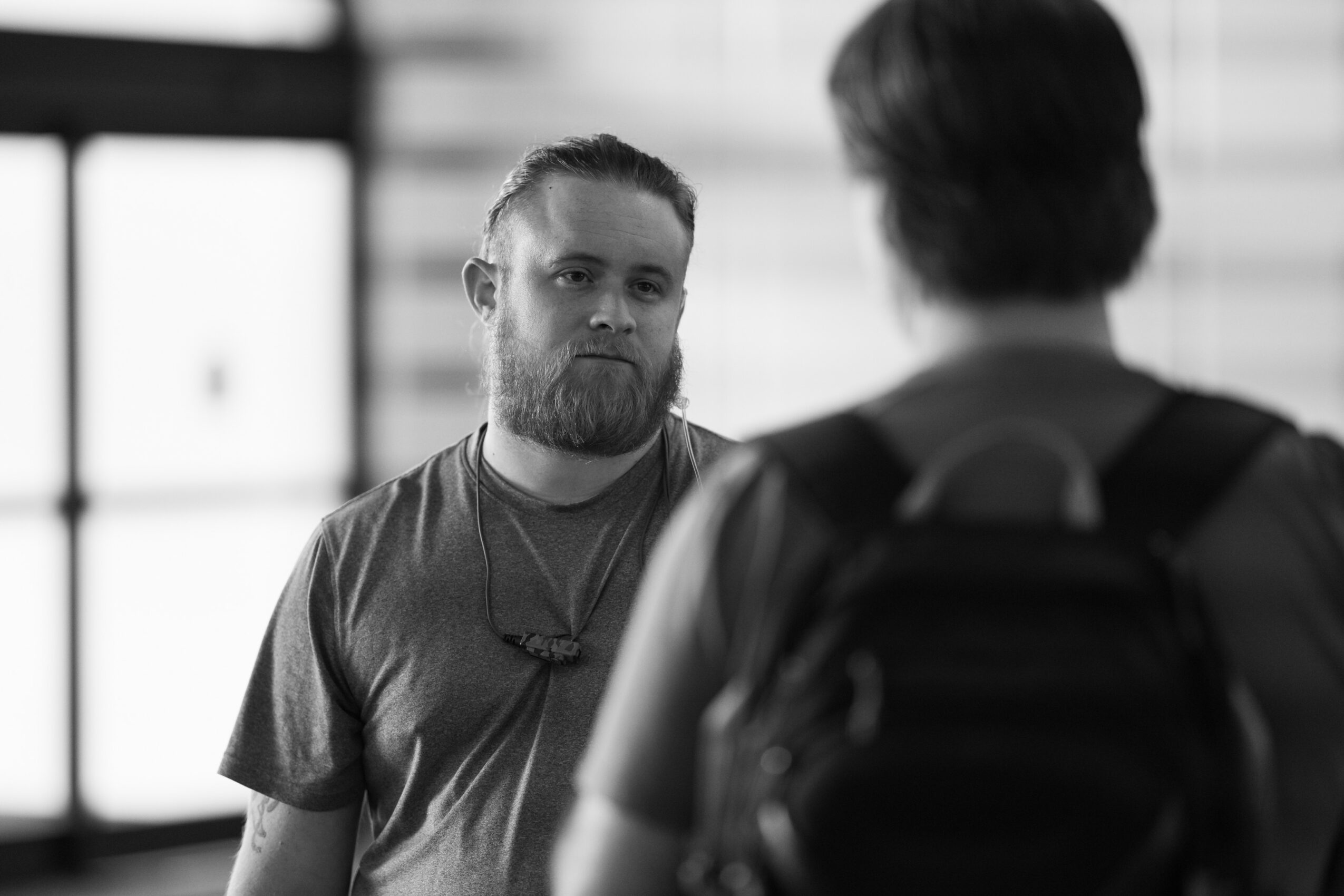 Two people talking indoors, one facing the camera with a beard and the other with their back to the camera, carrying a backpack.