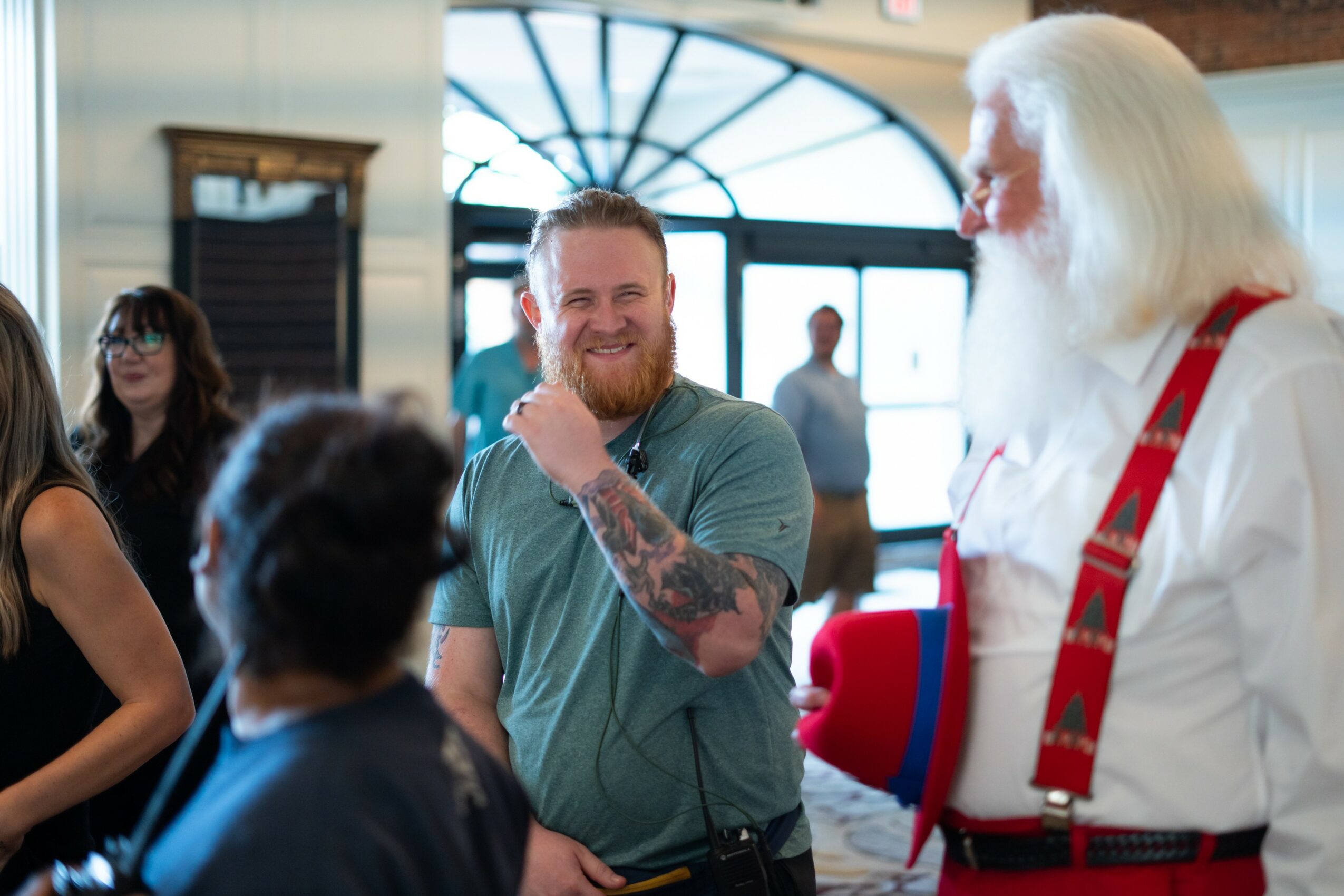 Group of people smiling and talking indoors, with a man with a beard and tattooed arm in focus.