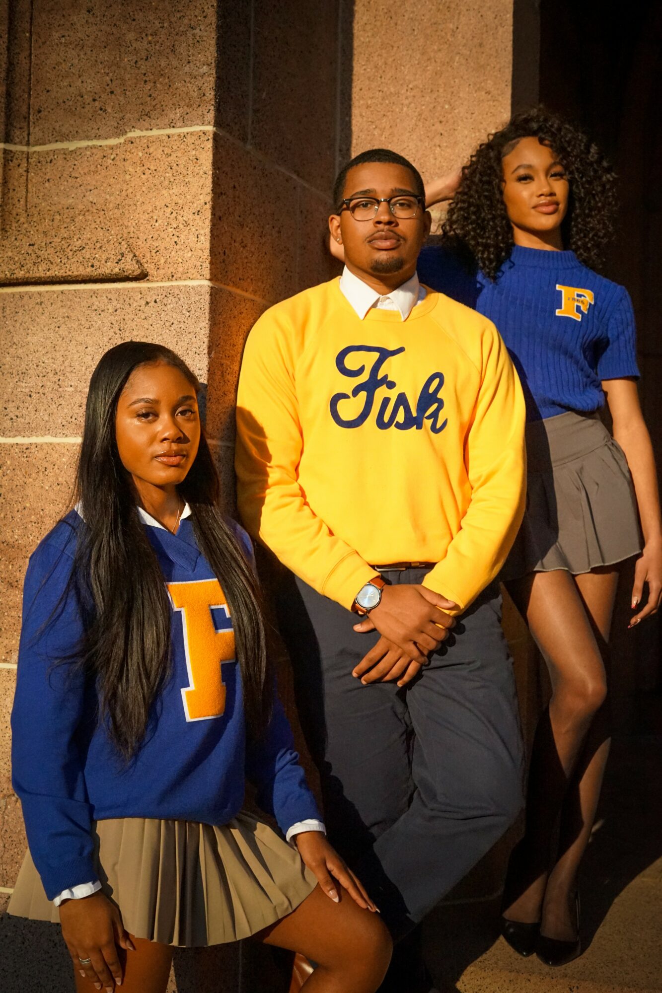 Three students in school uniforms standing against a brick wall, two women and one man, all looking forward.