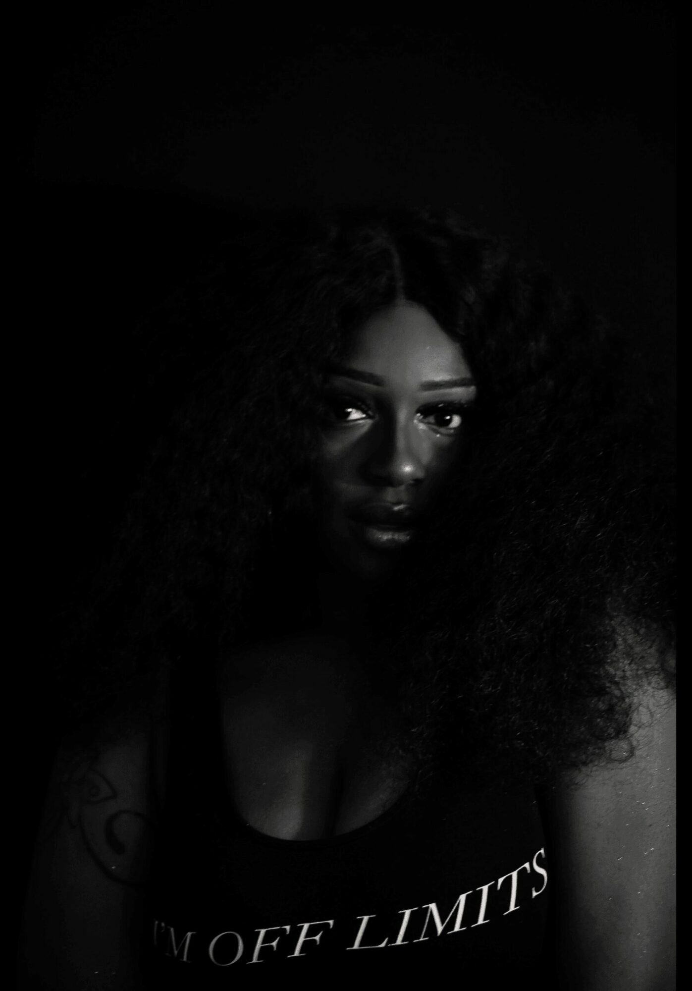 A woman with curly hair and dark clothing, partially illuminated, against a dark background.