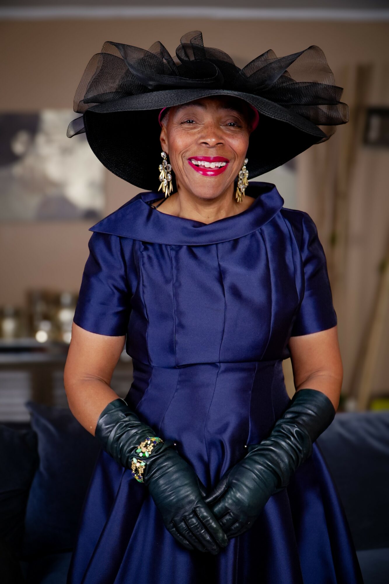 Woman in a blue dress and large black hat with tulle, smiling, wearing earrings and gloves, indoors.