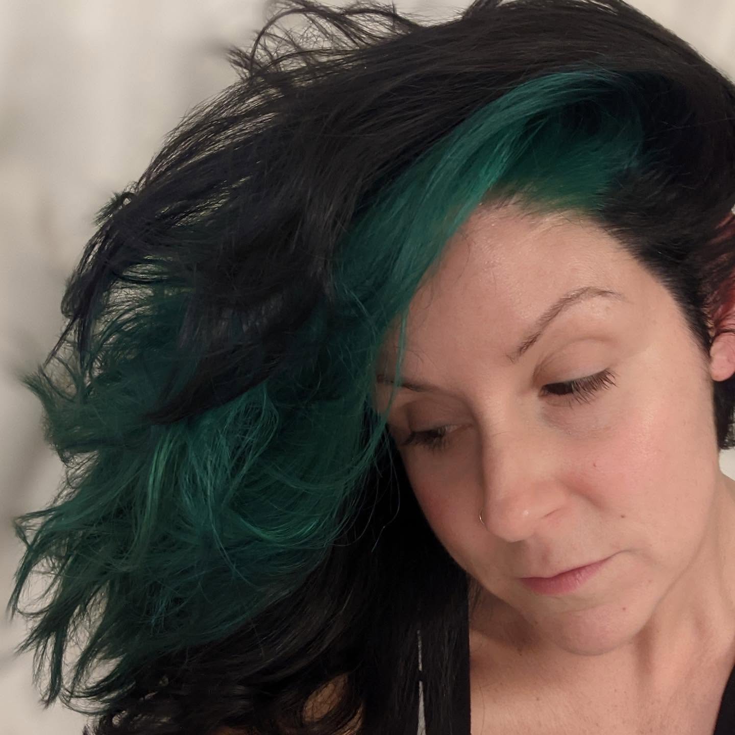 Close-up of a woman with black and green hair, looking down, with a neutral expression.