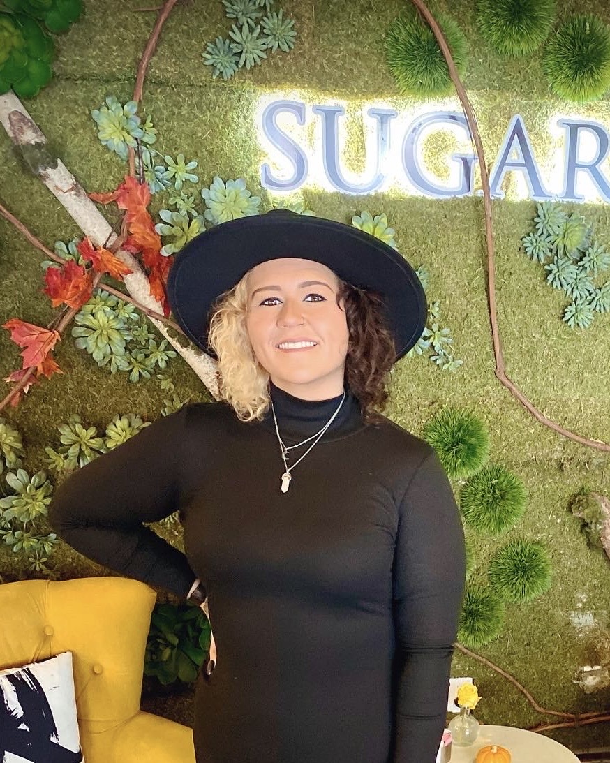 Woman with curly hair wearing a black hat and black top, smiling, standing in front of a green decorated background with the word 'SUGAR'.