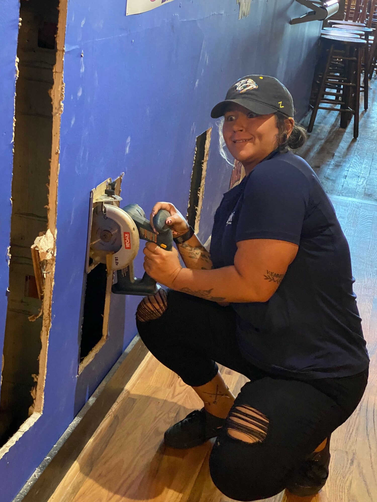 Person wearing a helmet and black clothing using a circular saw on a wooden floor, with a blue wall in the background.