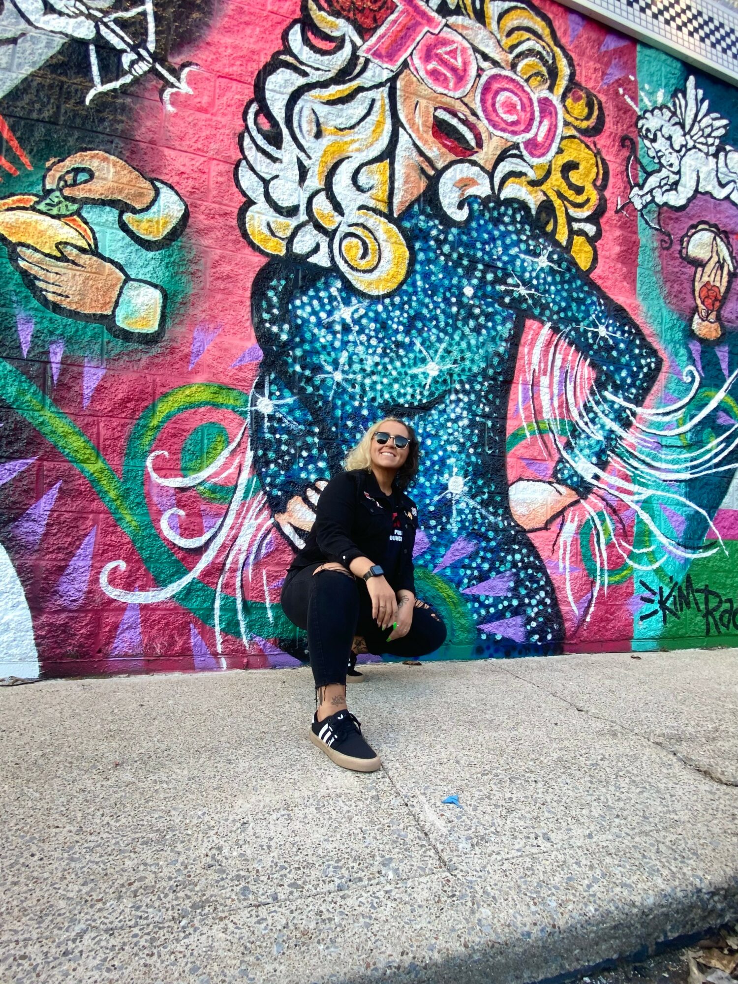Person sitting in front of a colorful mural of a woman with curly hair, sunglasses, and a starry dress, smiling.