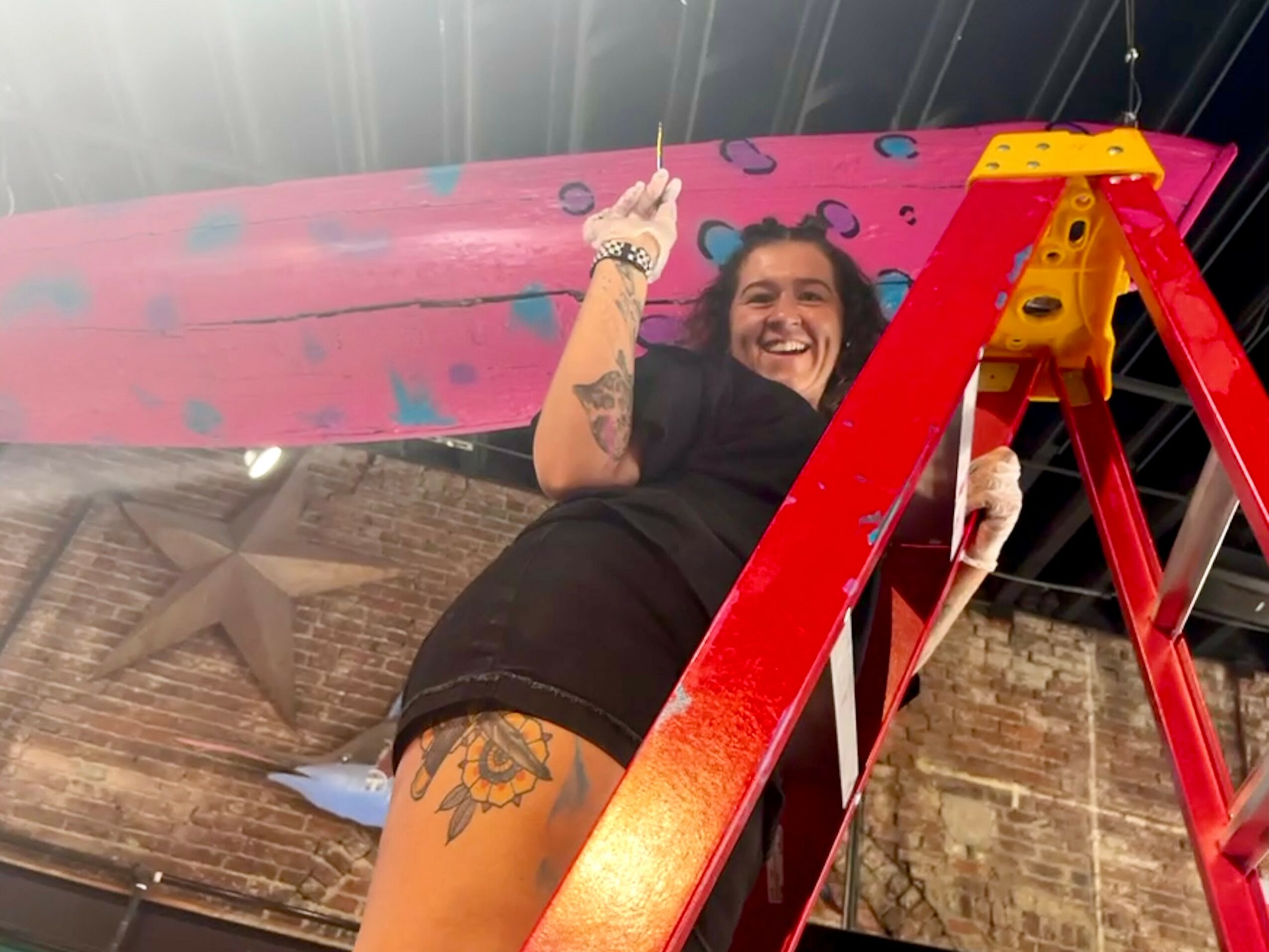 Woman smiling on a pink skateboard, standing on a red ladder, with a brick wall background.