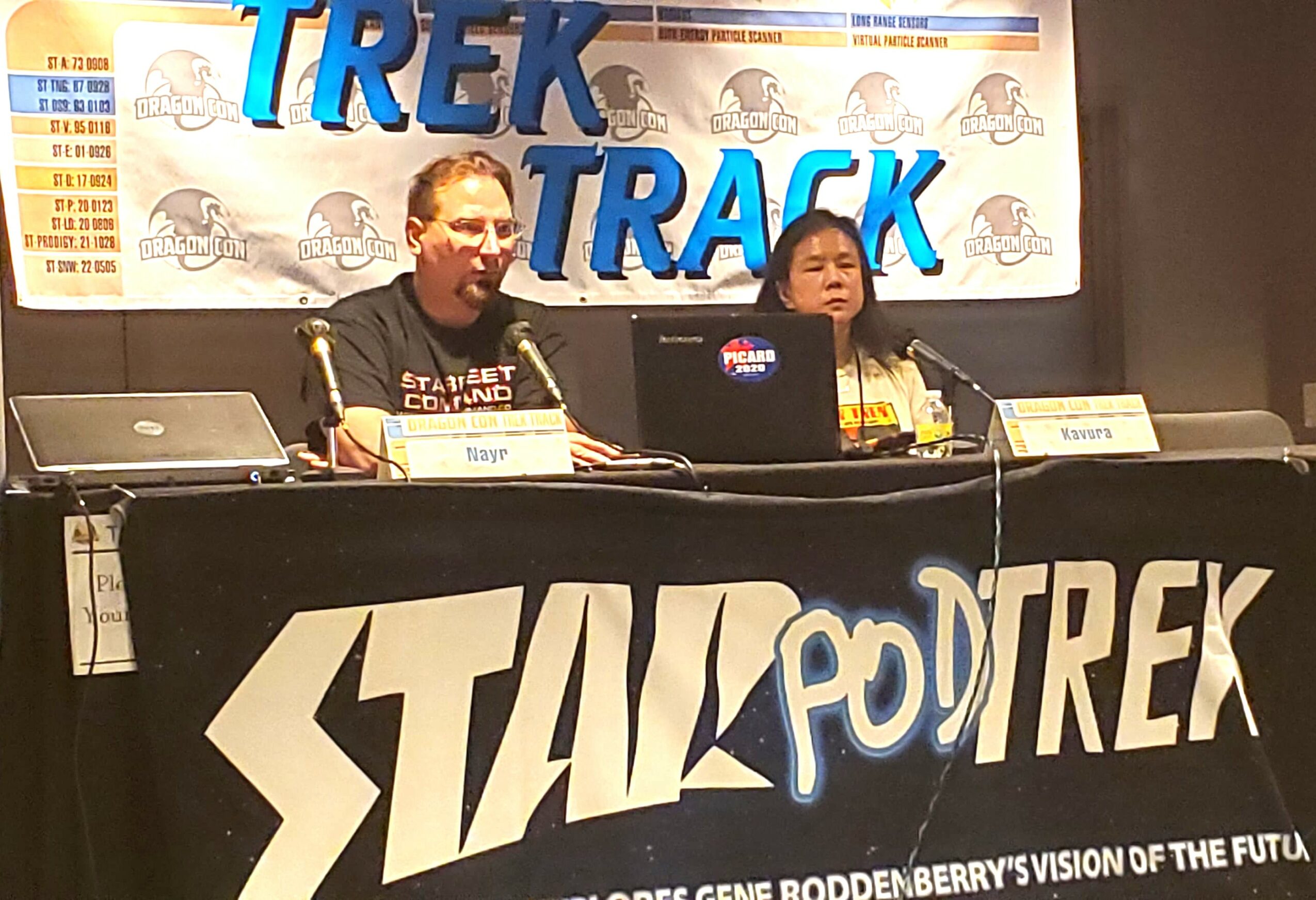 Two people sit at a table with a Trek Track banner behind them, speaking into microphones at a conference or event.