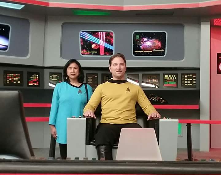 Two people in Star Trek costumes on a spaceship bridge set with control panels and screens behind them.