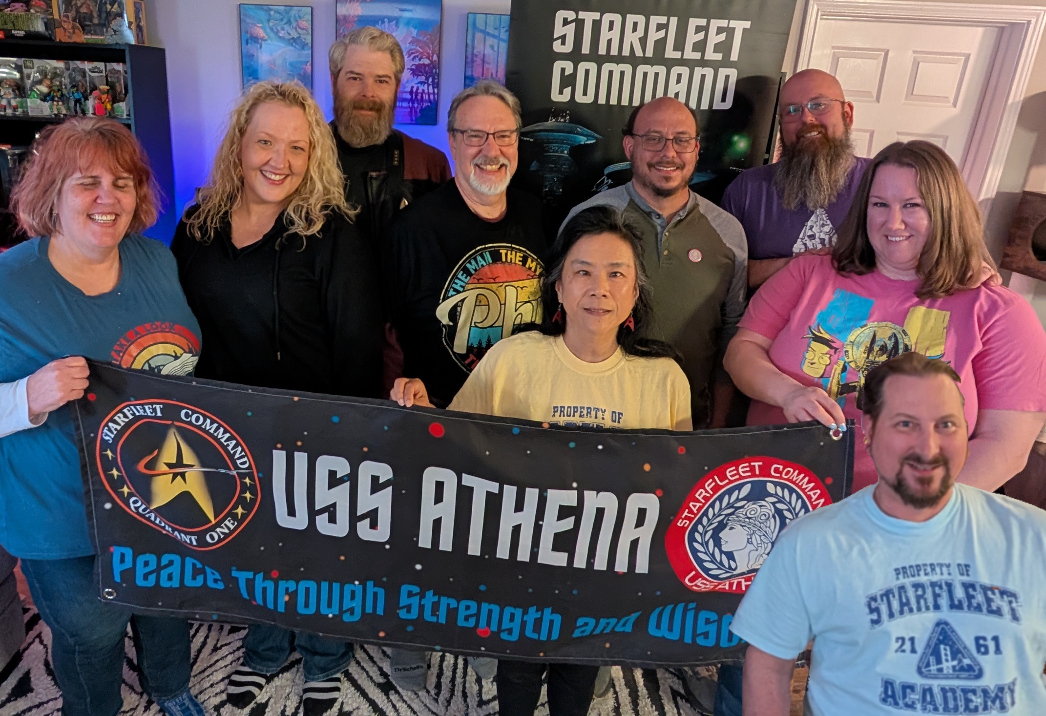 Group of nine people holding a USS Athena banner, smiling indoors with a Starfleet Command poster in the background.