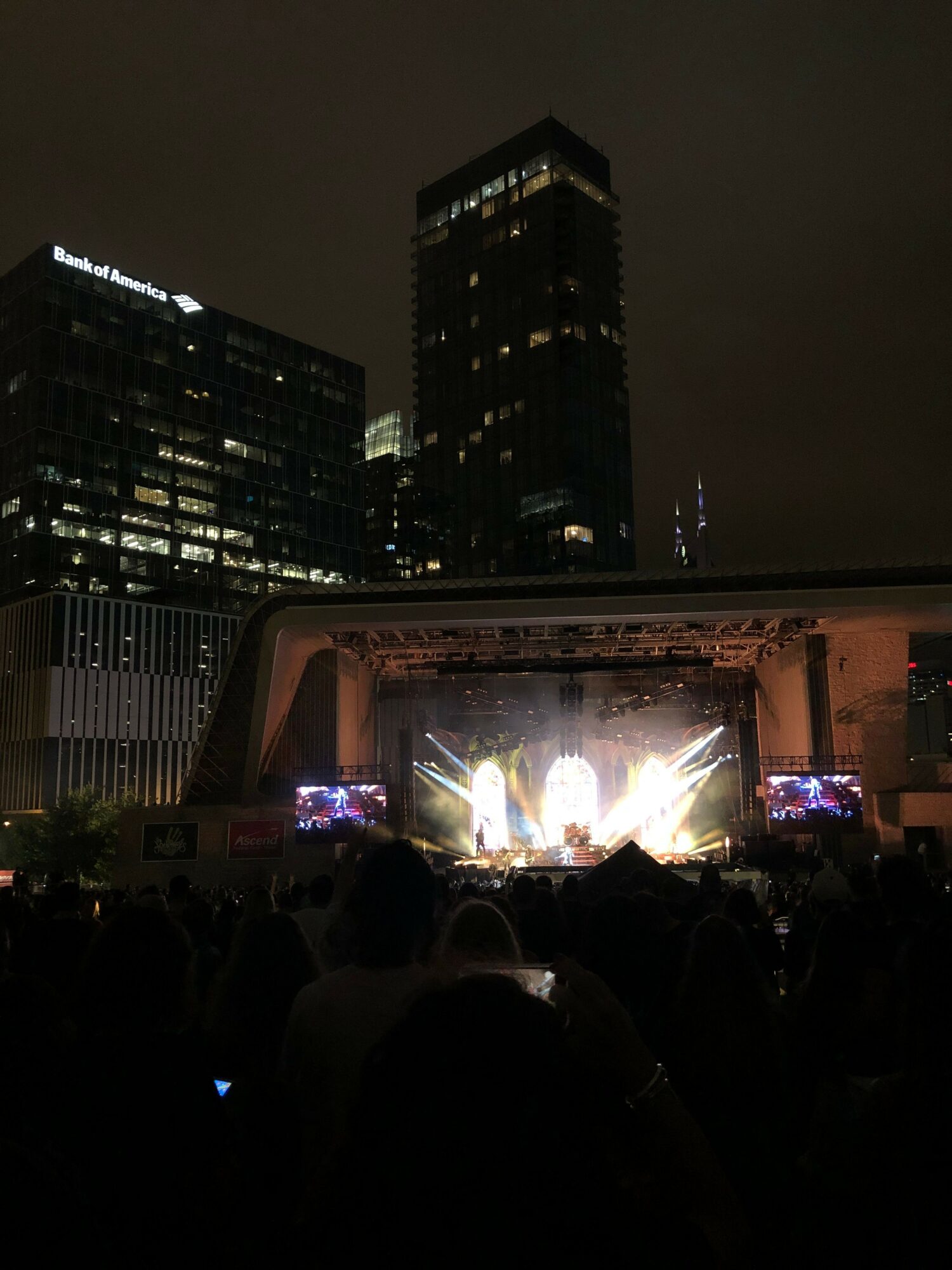 Nighttime outdoor concert with a stage, bright lights, and an audience, in front of tall buildings.