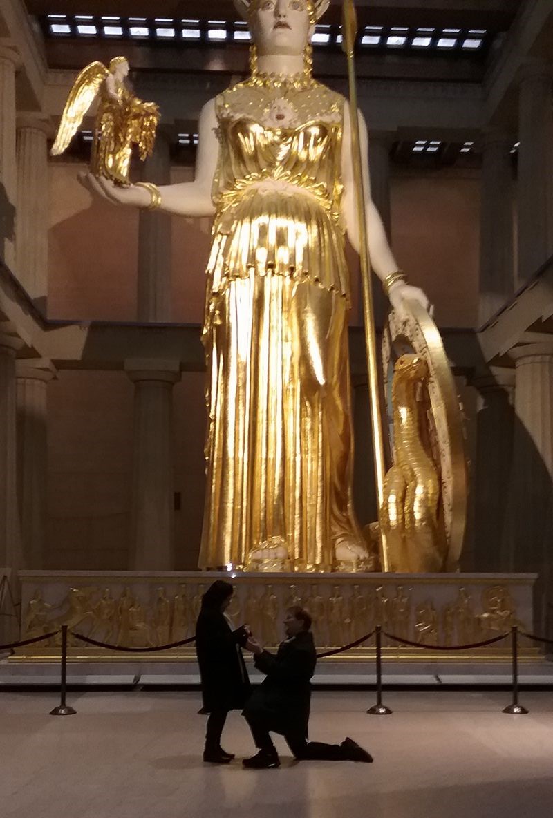 Large golden statue of a woman holding a bird and a shield, with two people kneeling in front, in a museum setting.