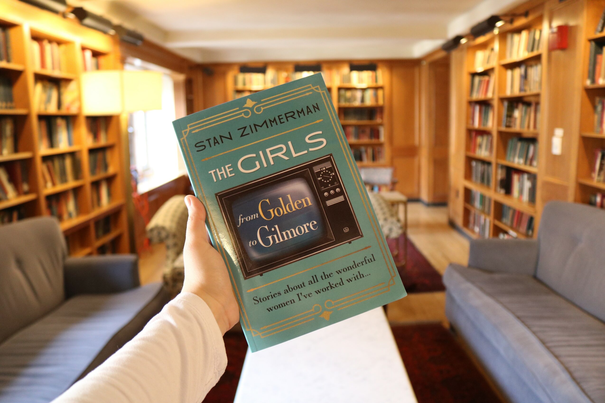 Person holding a book titled 'The Girls' by Stan Zimmerahn in a library with bookshelves and sofas.