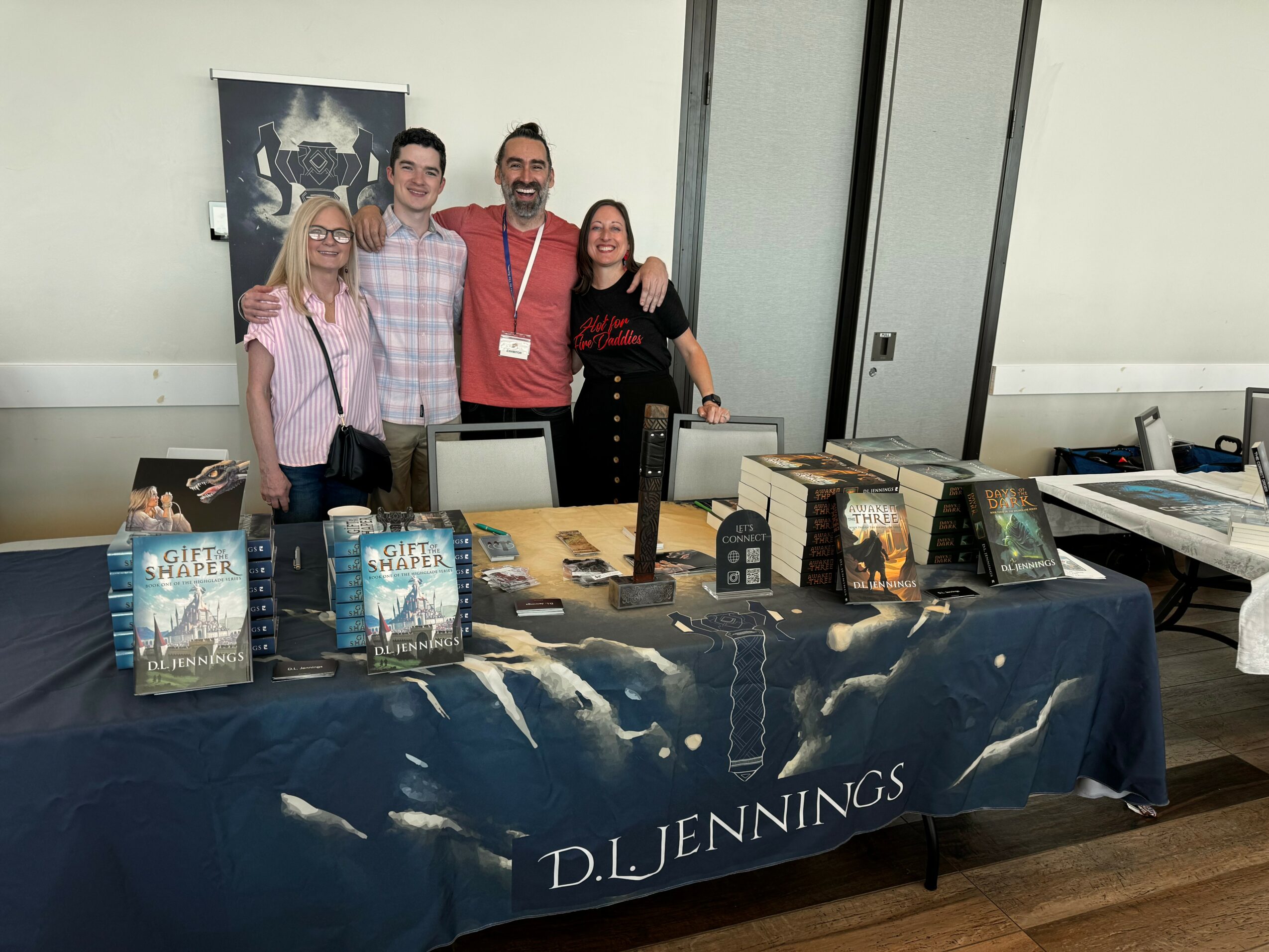 Four people stand behind a table with books and merchandise, smiling at the camera, in a room with white walls.