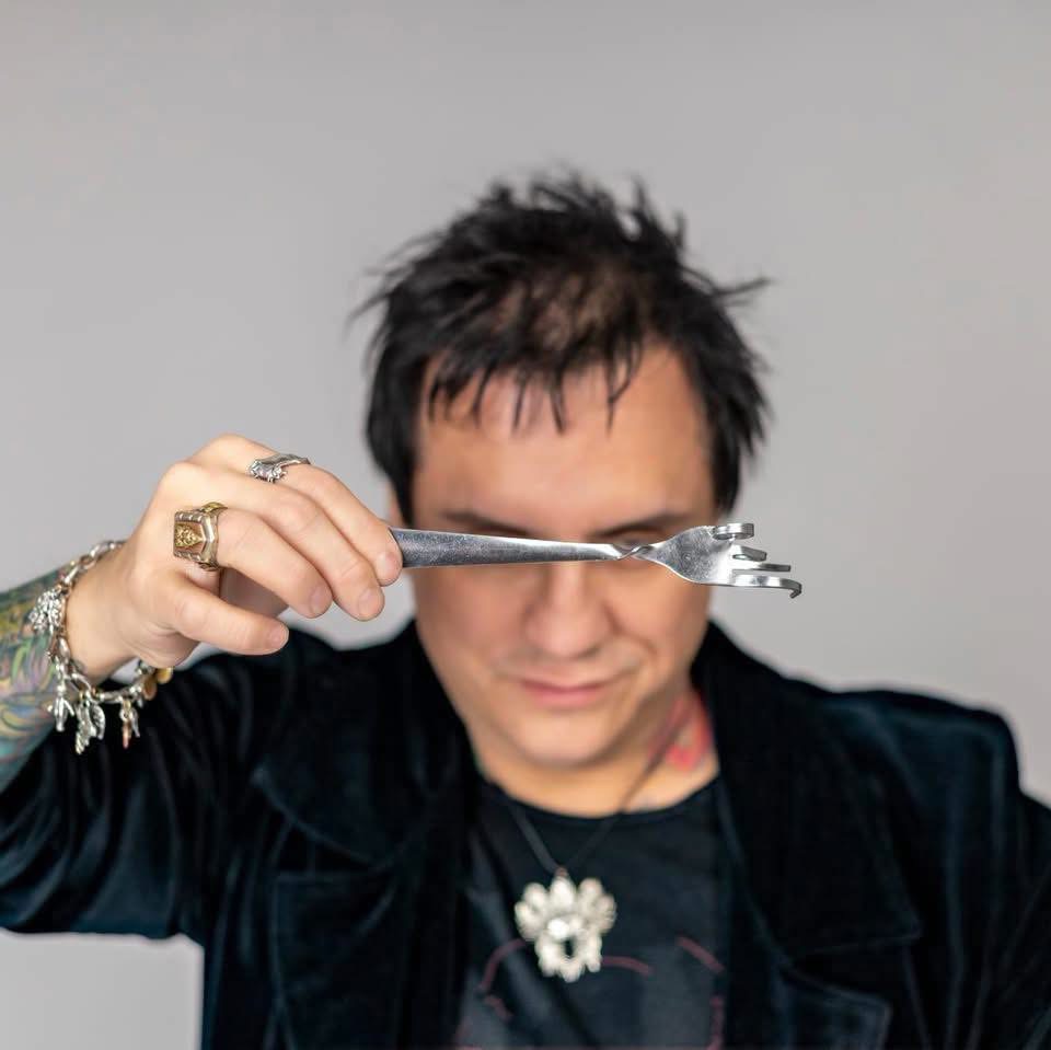 Person with dark, spiky hair holds a silver object near their eye, wearing rings and a necklace with a flower pendant.