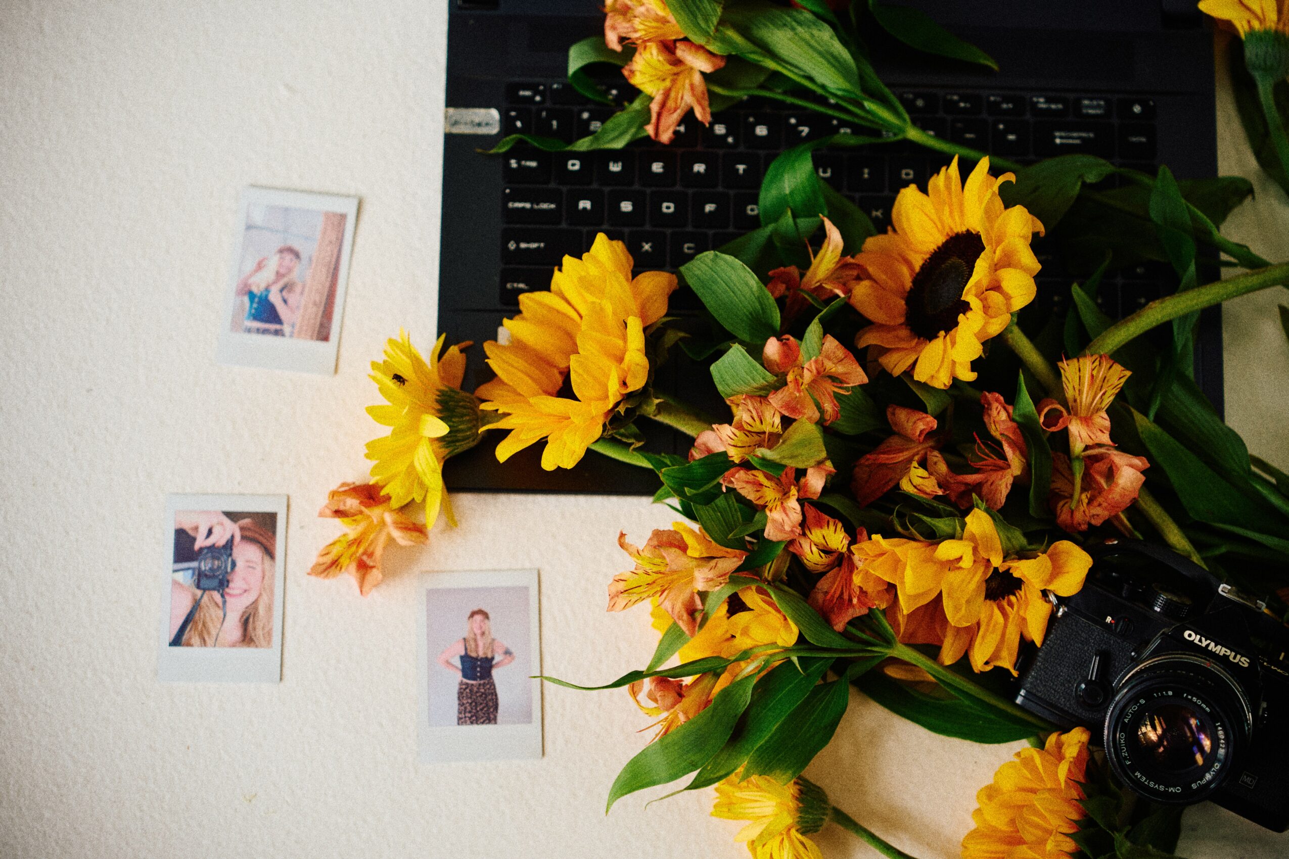 A bouquet of sunflowers and orange flowers on a desk with a camera, with small photos on the wall behind.
