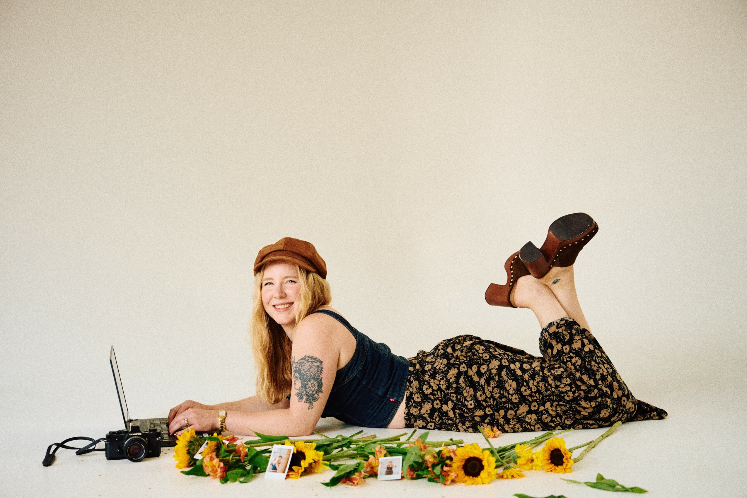 Woman lying on her stomach on a blanket with sunflowers, smiling, wearing a hat and floral pants, holding a camera.