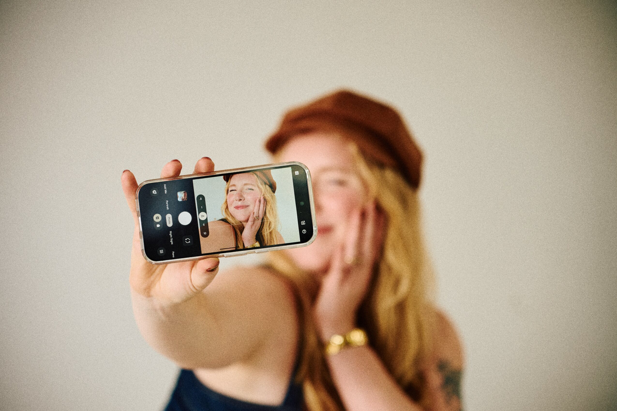 Person with long hair and a hat takes a selfie with a smartphone, smiling and holding the phone in front of face.