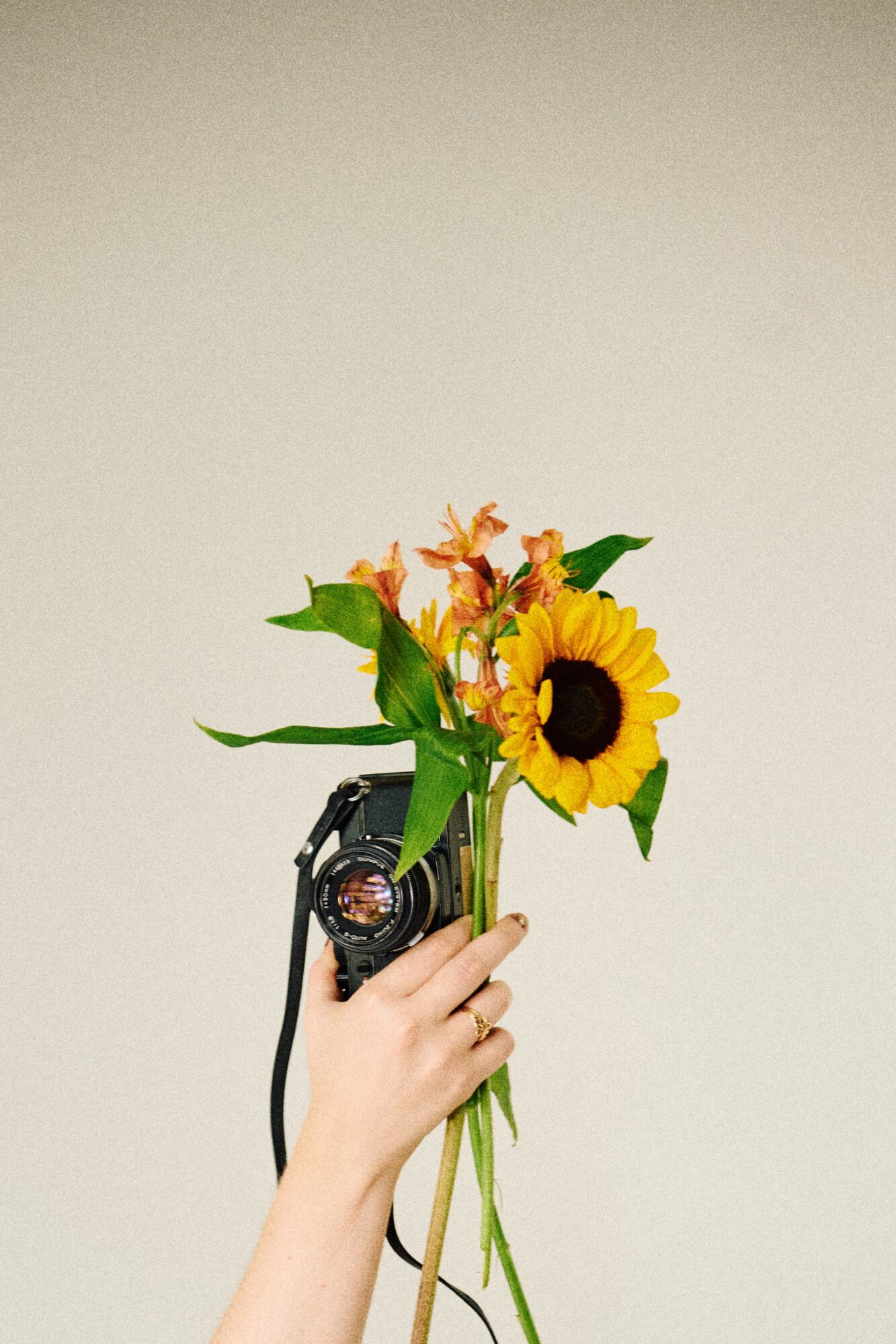 Hand holding a camera with a bouquet of sunflowers and orange flowers against a plain background.
