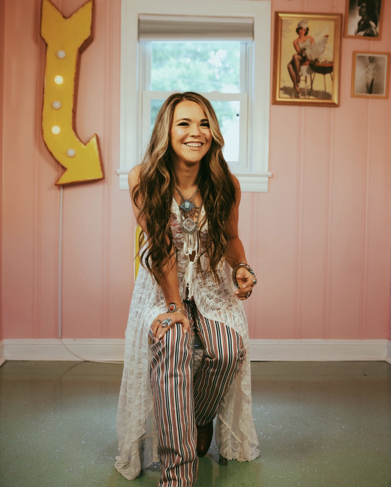 Young woman with long hair smiling, sitting on a stool in a pink room with framed pictures and a large yellow letter decoration.
