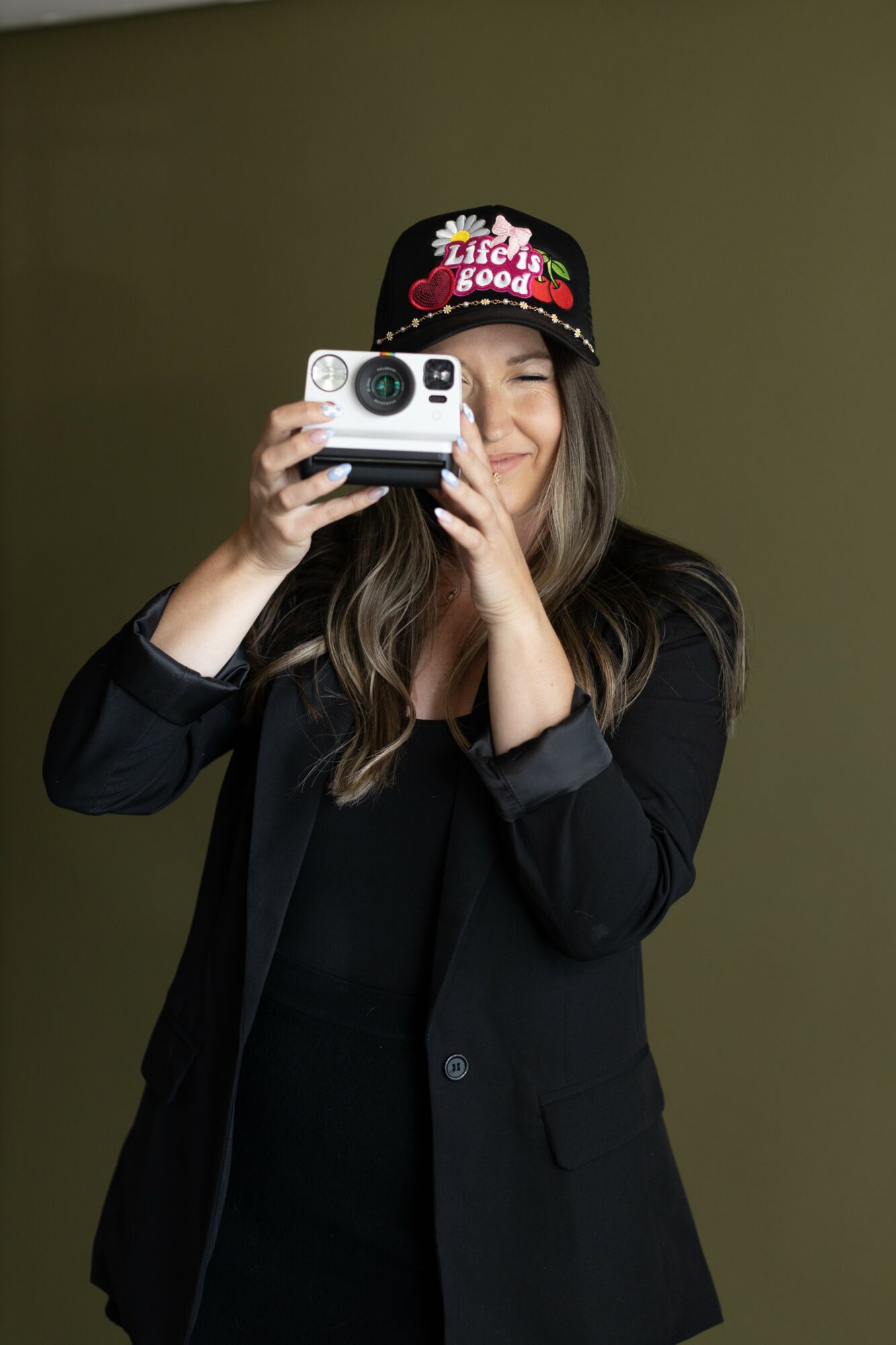Woman taking a photo with a camera, wearing a black blazer and a black cap with colorful text and graphics, smiling.