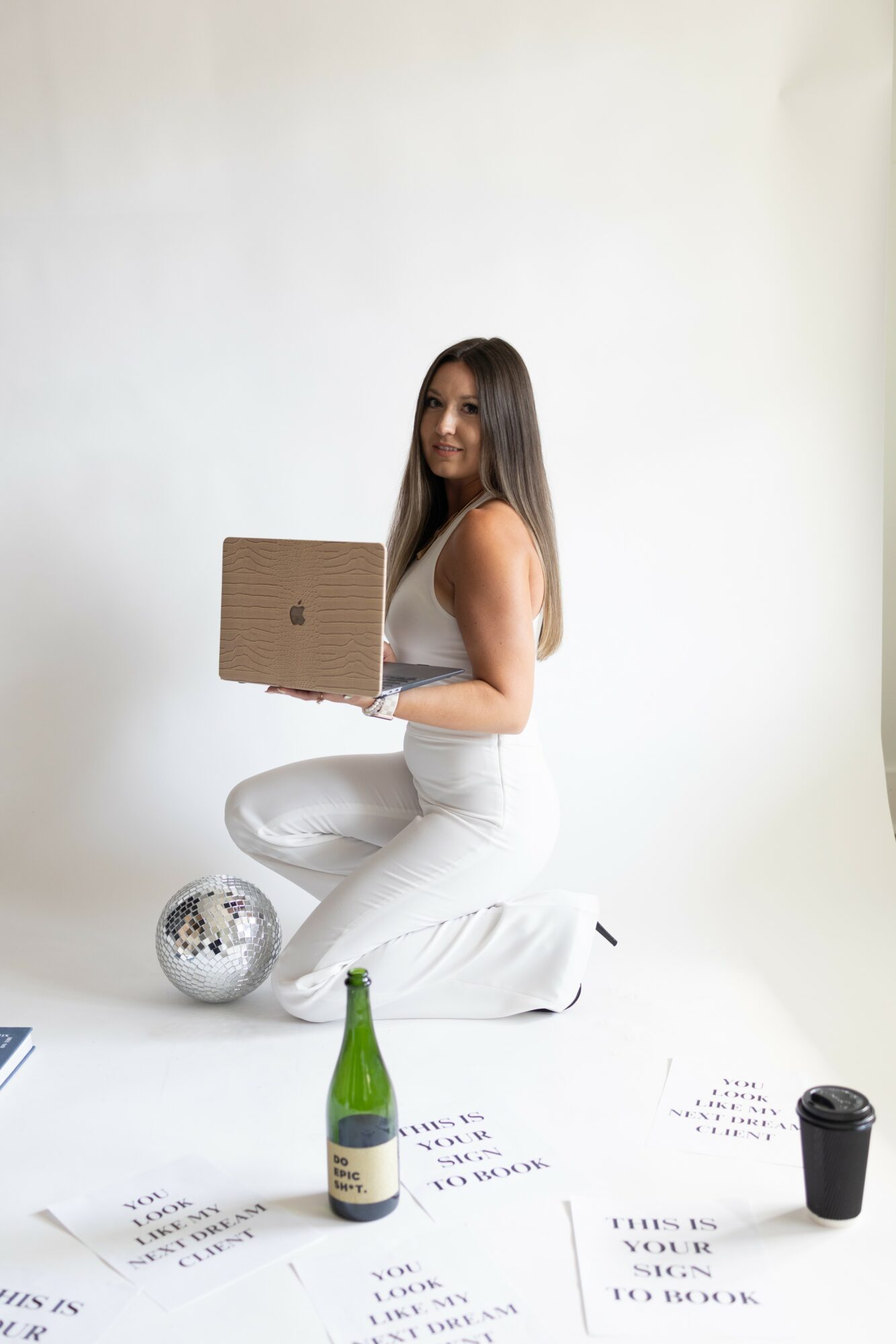 Woman kneeling on floor holding a cardboard box, surrounded by a green bottle, papers, and a disco ball, against a plain white background.