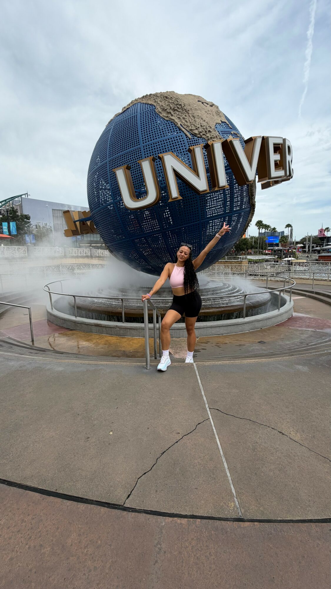 Person standing in front of the Universal Studios globe fountain, arms raised, outdoors on a cloudy day.
