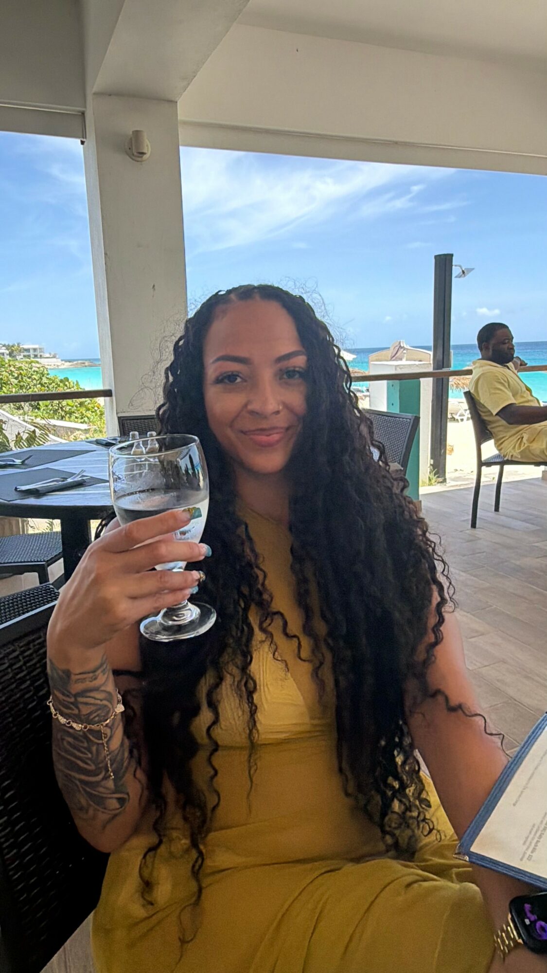 Woman with long curly hair holding a glass, sitting outdoors with a blue sky and ocean in the background.