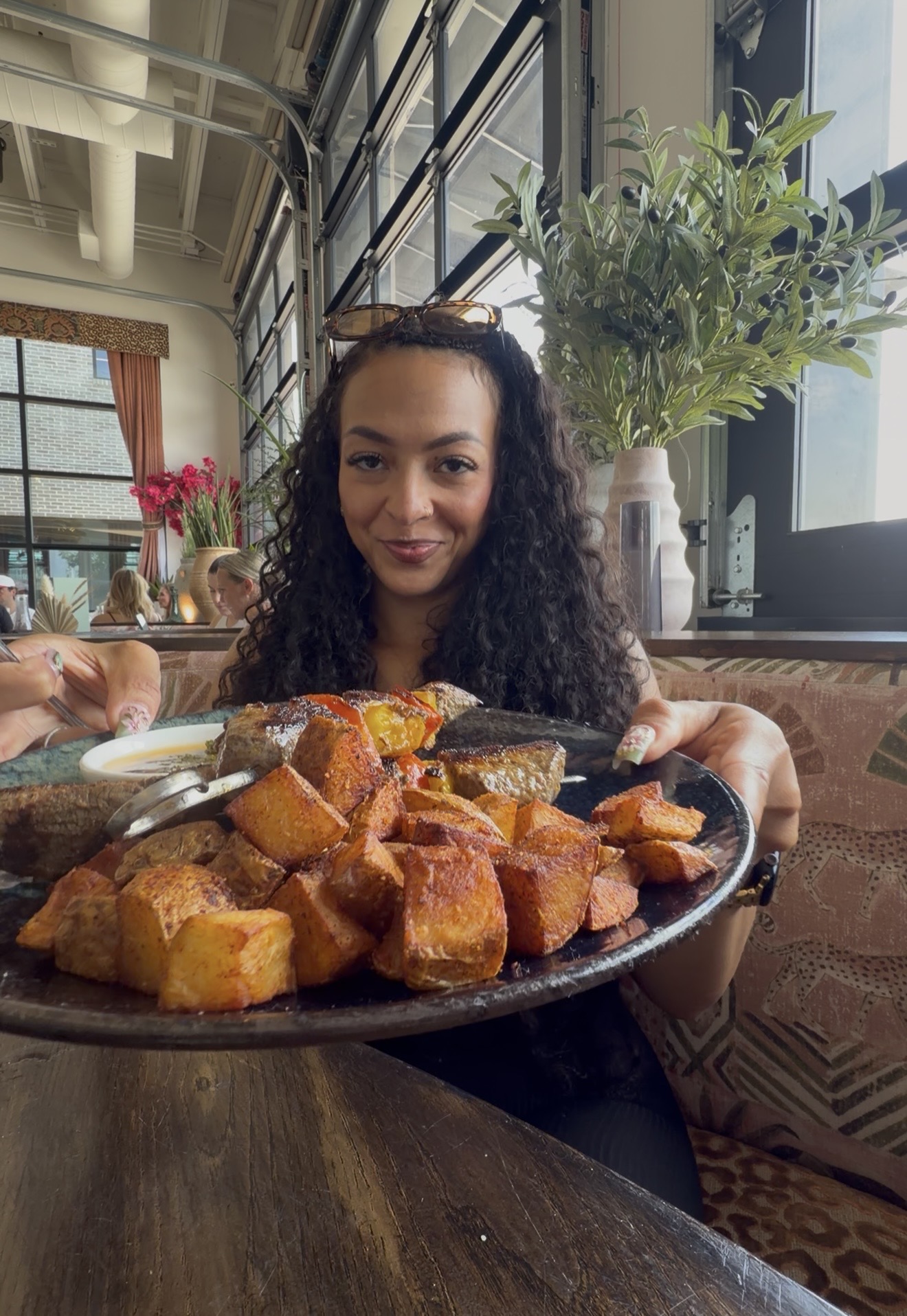 Young woman with curly hair holding a plate of fried food in a restaurant with large windows and plants.
