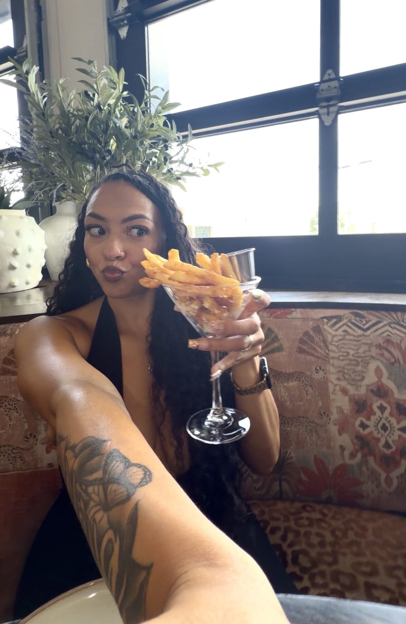 Woman with curly hair holding a glass of fries, sitting in a restaurant with large windows and plants behind her.