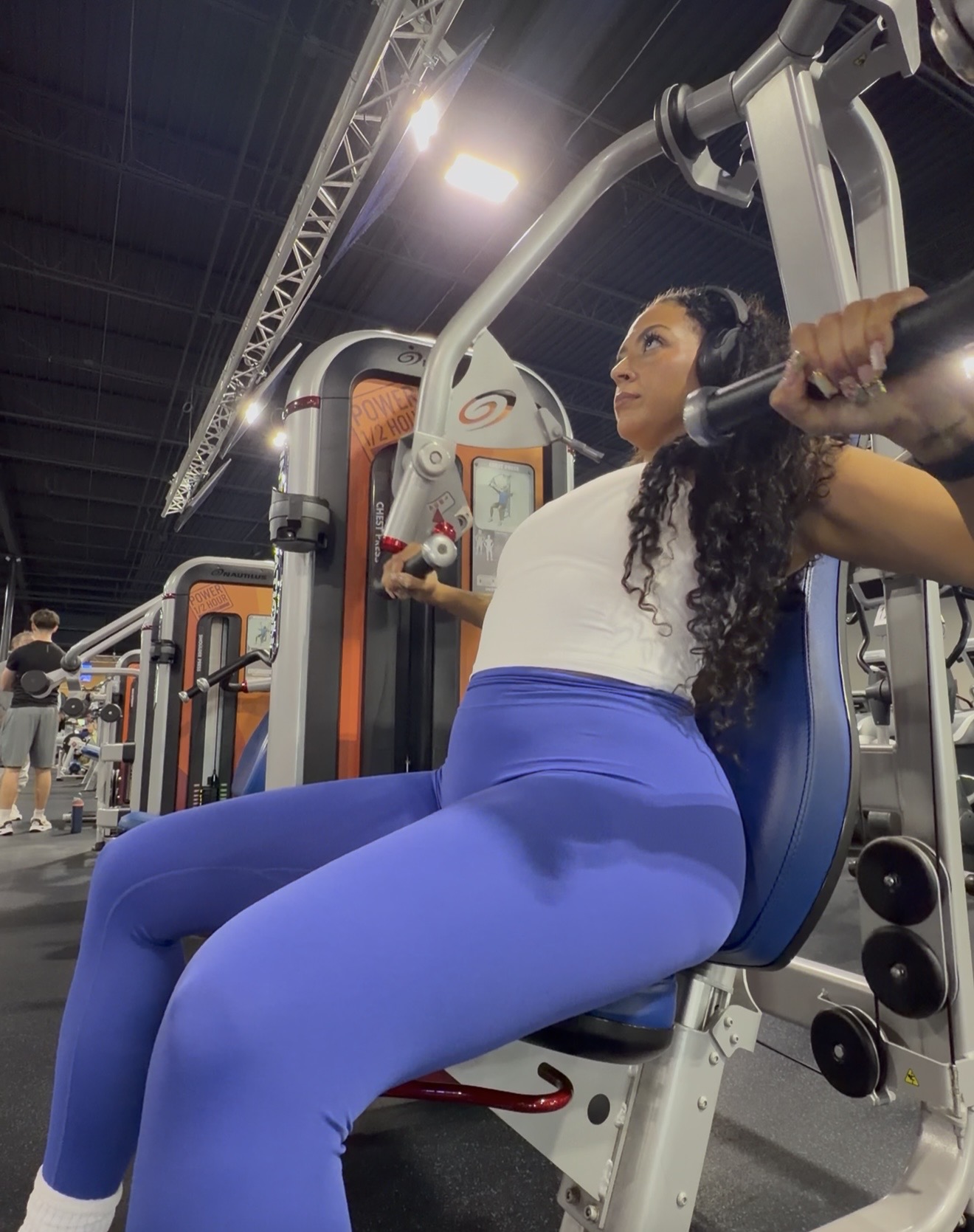Woman using a gym machine, wearing a white top and blue leggings, in a fitness center with equipment and lighting.