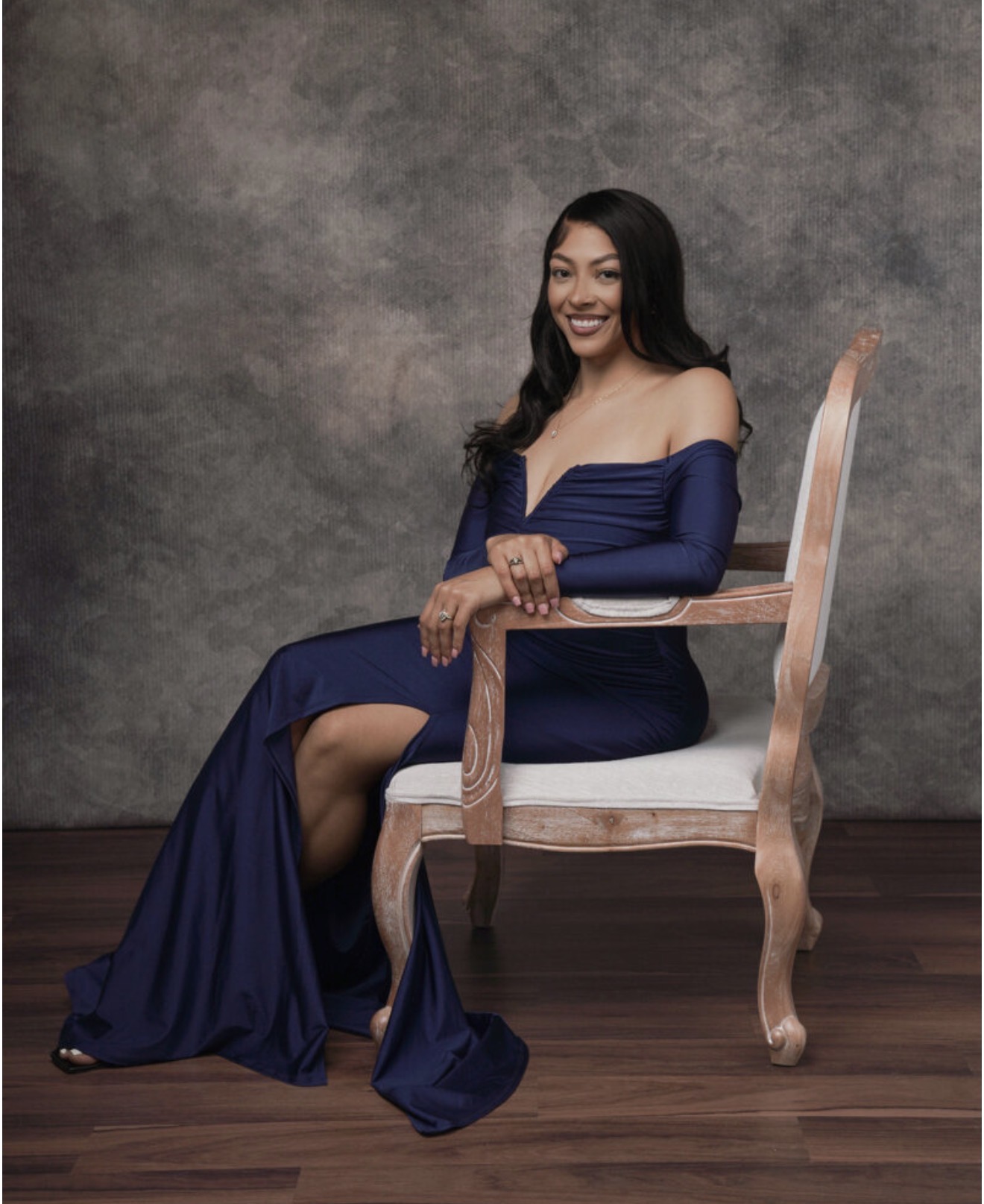 Woman in a blue off-shoulder gown sitting on a wooden chair, smiling, against a textured gray background.