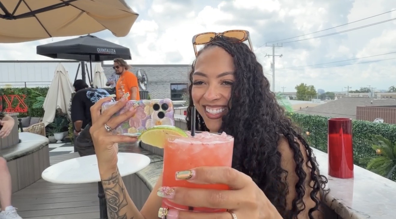 Woman with curly hair smiling and holding a pink drink outdoors, sunglasses on head, with a patio and sky in background.