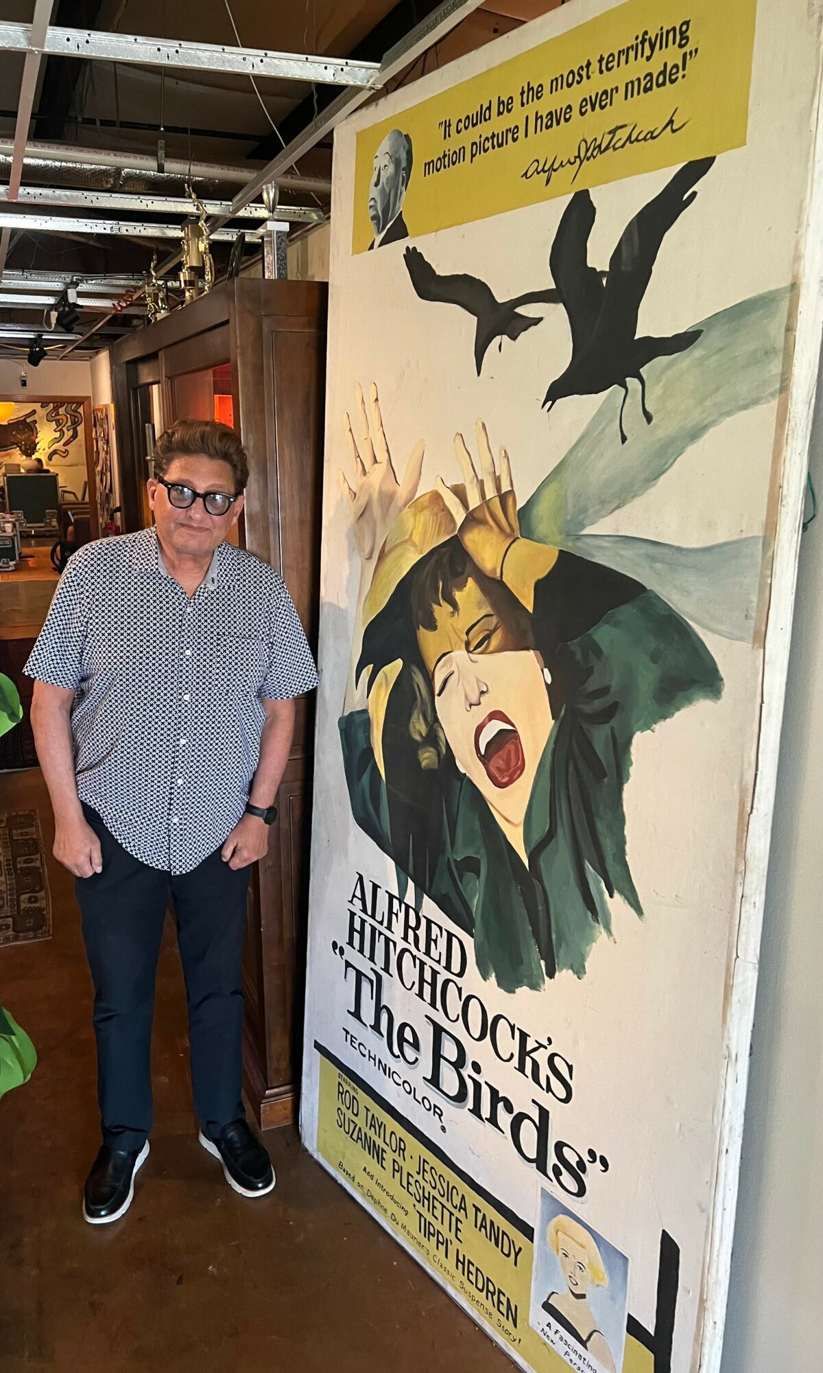 Person with glasses and short hair standing next to a poster for the play 'The Birds' with a woman screaming and a bird flying overhead.