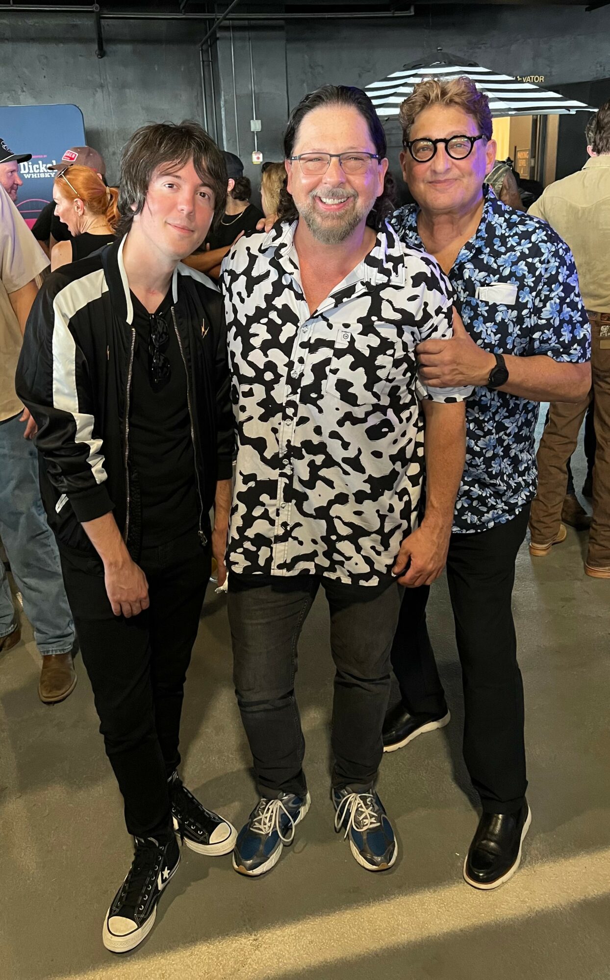Three people standing together indoors, smiling, with a crowd in the background.
