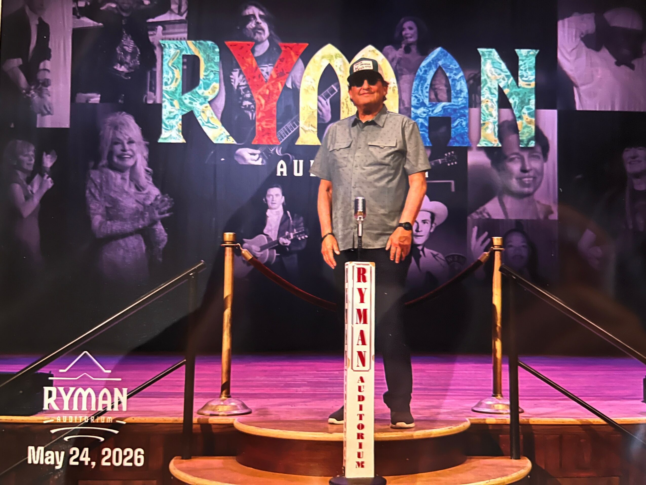 Person standing on stage with a sign, in front of a colorful backdrop with the word 'RYMAN' and photos, on a wooden stage.