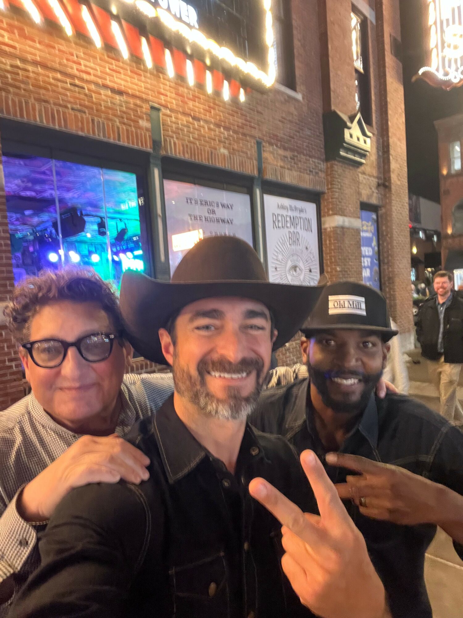 Three people posing indoors, one wearing a large hat, two with glasses, making peace signs, brick wall background.
